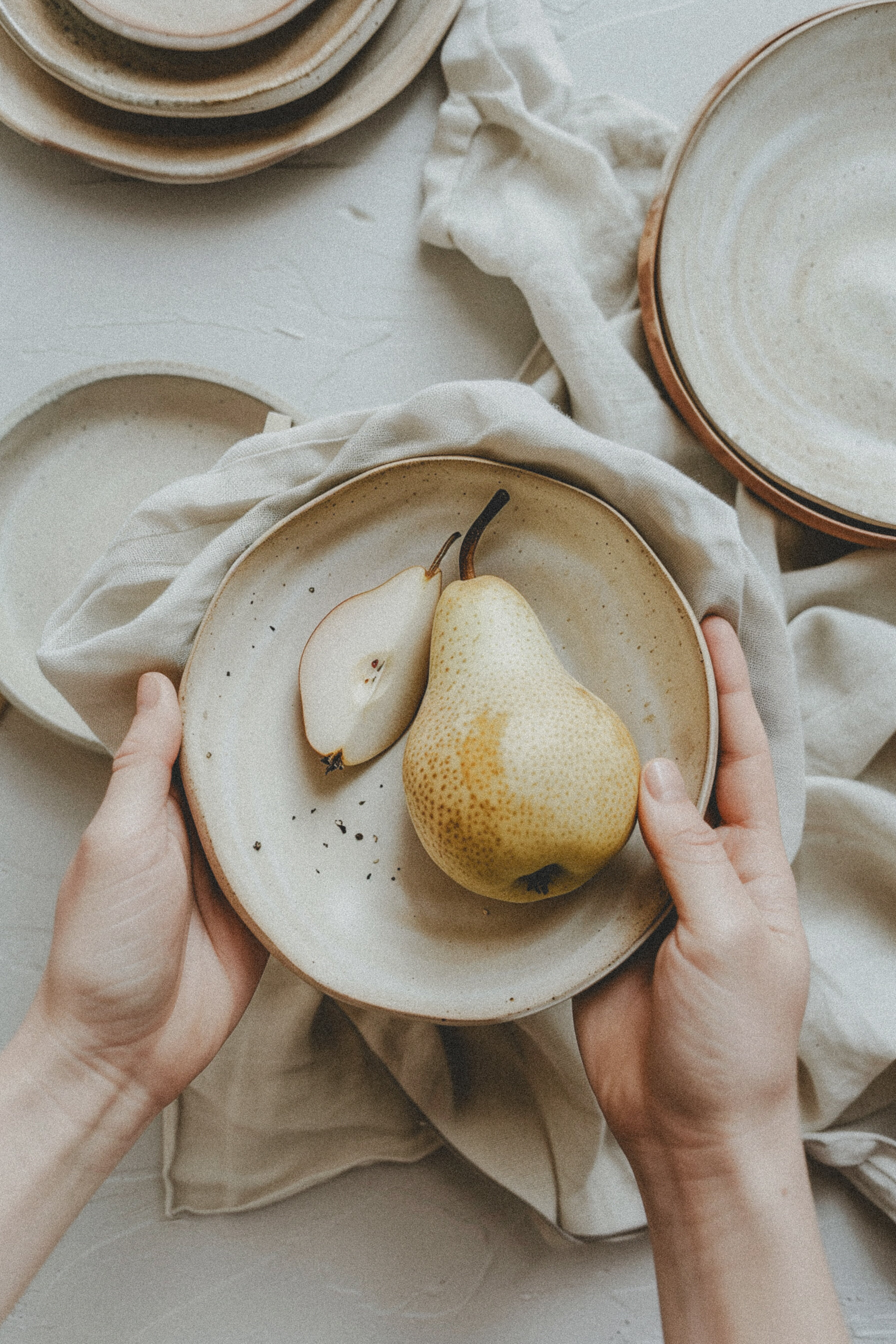two hands holding a pottery  plate with a cut pear over a pile of linen cloth and other pottery plates