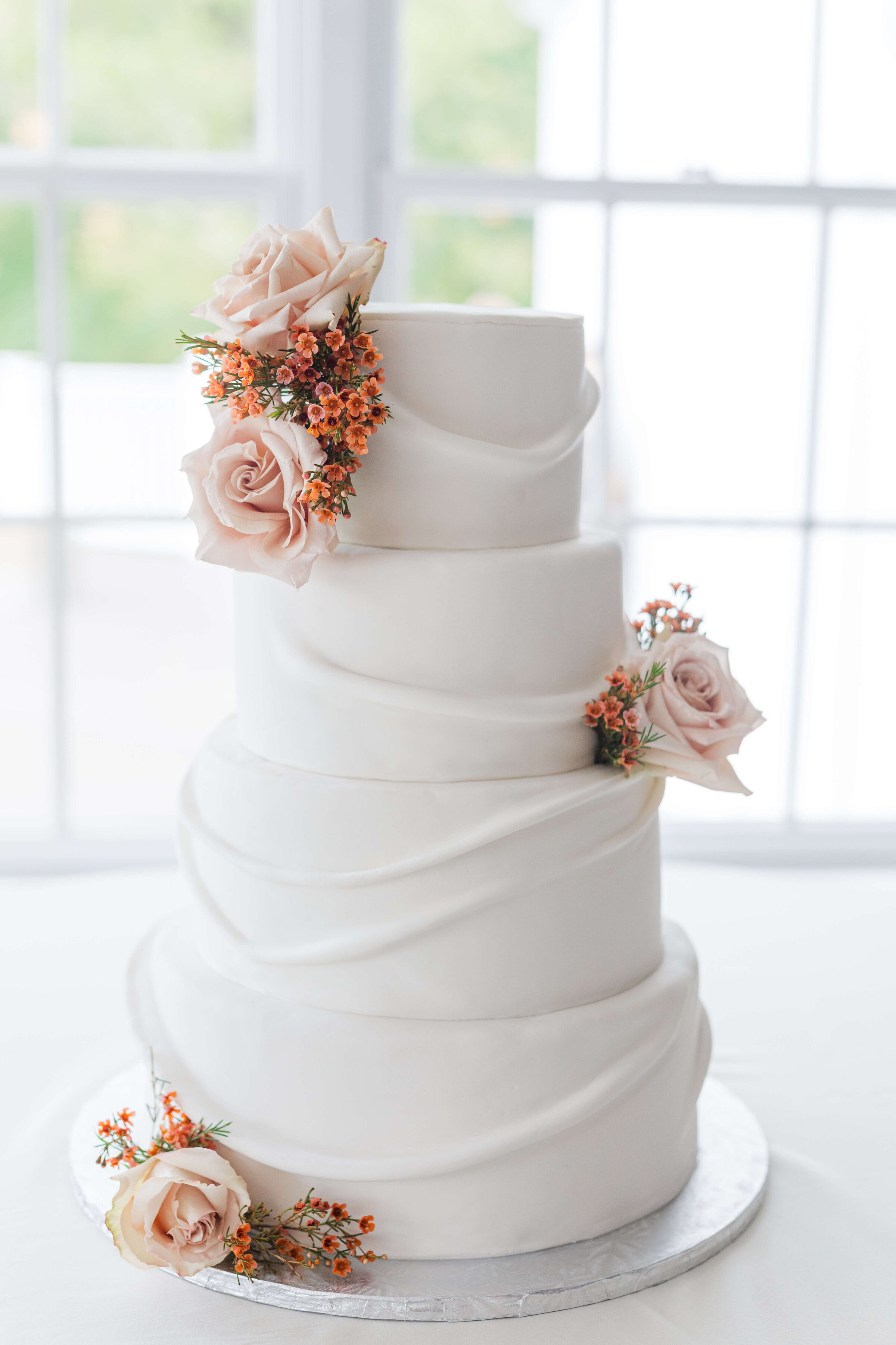 A white wedding cake with peach flowers on it. It's in front of a white window and on top of a white table cloth