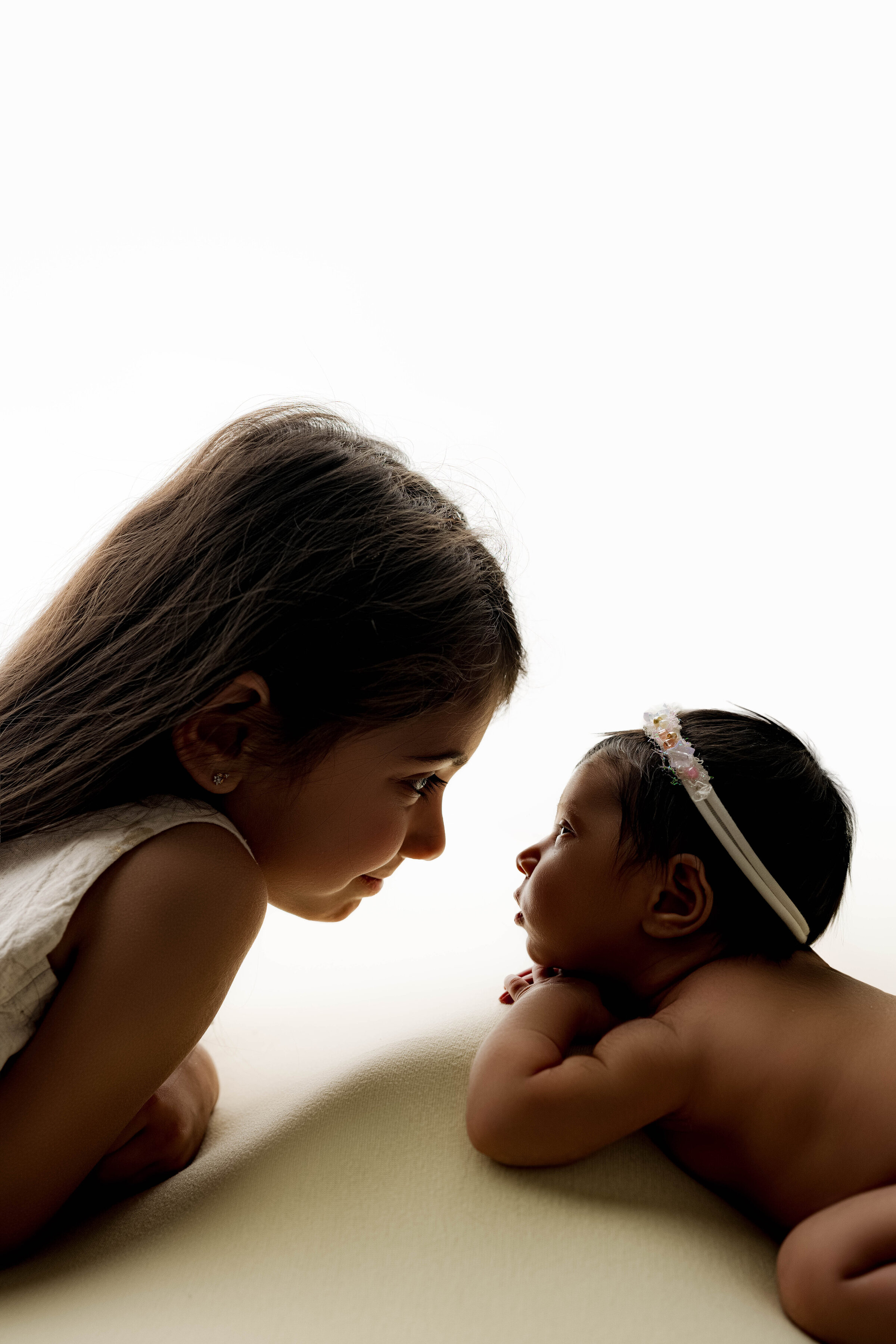 Older sister and newborn baby gazing at each other, lying face-to-face on a light yellow backdrop.
