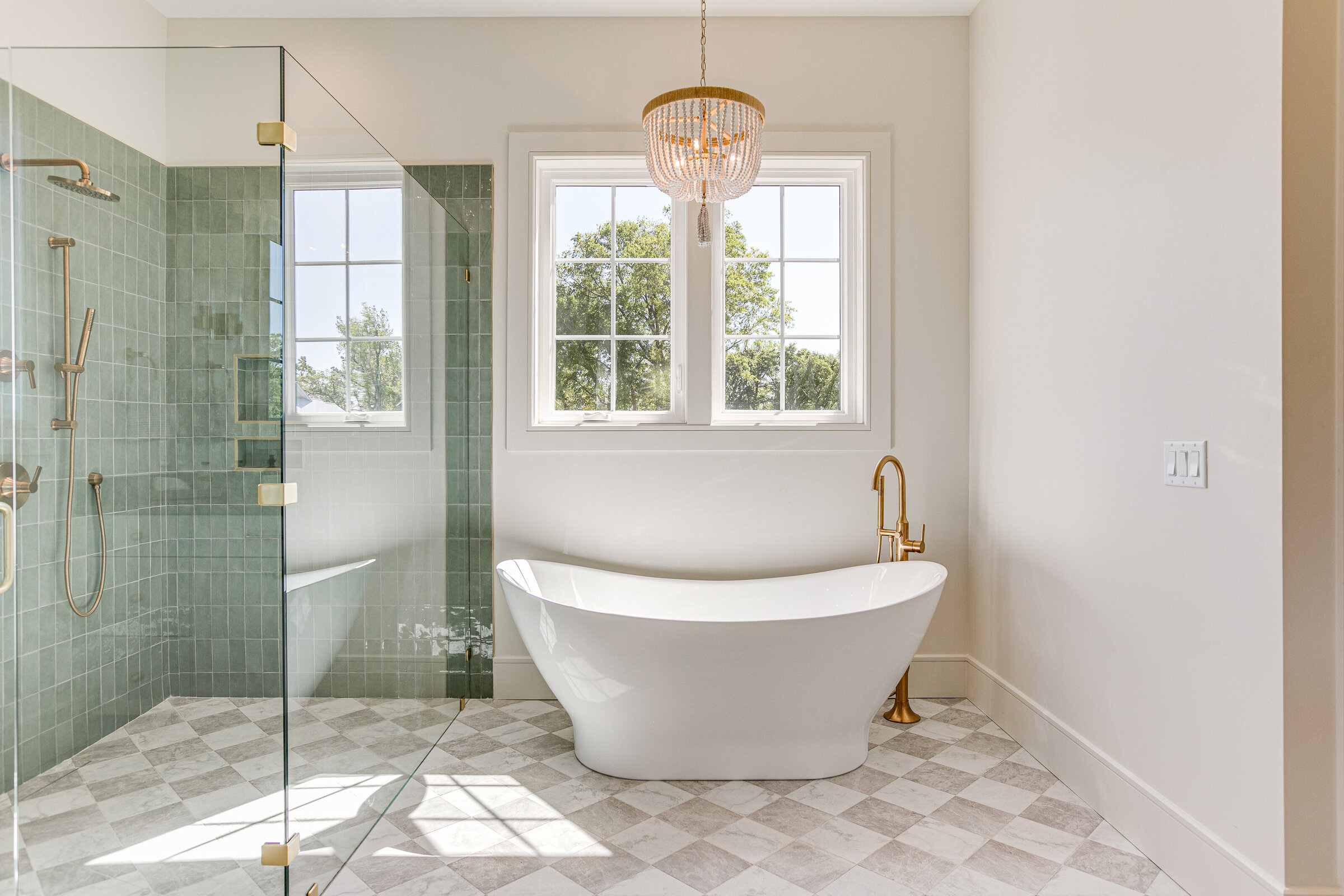 A stunning editorial shot featuring a sage green tile shower, a large soaking tub, and elegant glass windows. The space is accented by a coastal chandelier and gold finishes, creating a luxurious and serene bathroom environment.