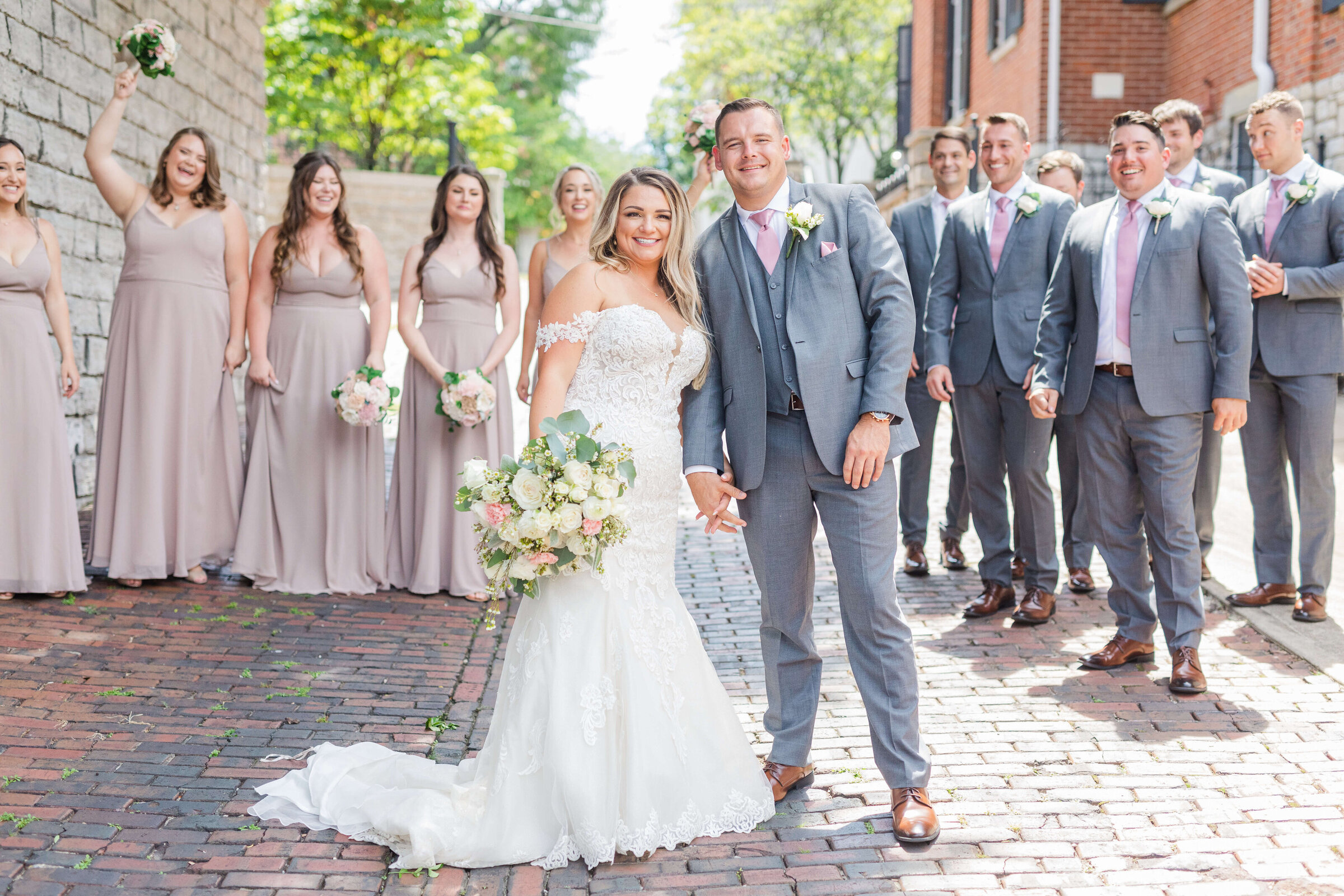 A bride and groom smile at the camera while their bridal party walks in back of them. They are on cobblestone in the summer time. The bridesmaids are wearing taupe and the groomsmen in grey.