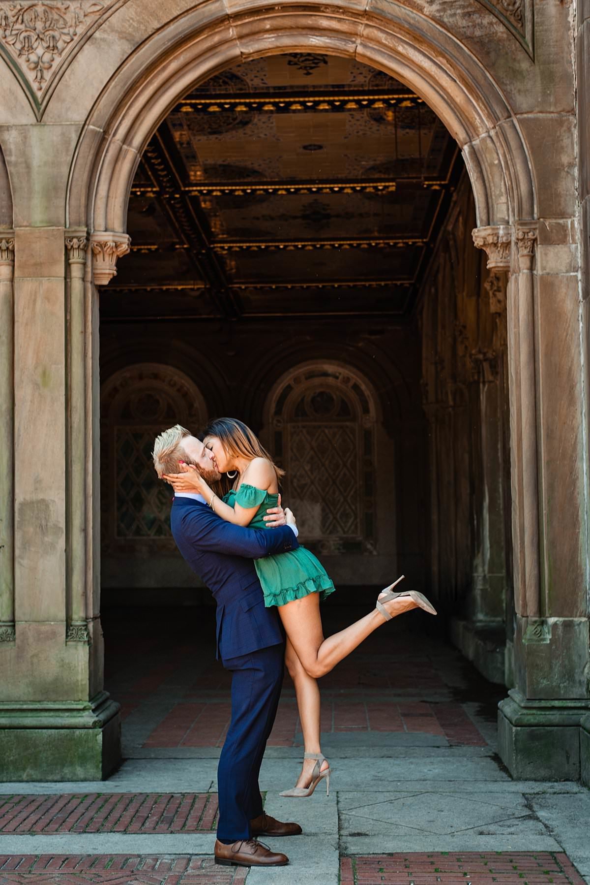 Man lifting his girlfriend for a heel popping kiss under iconic Central Park archway in New York, he is in a blue suit and she has on a short green dress
