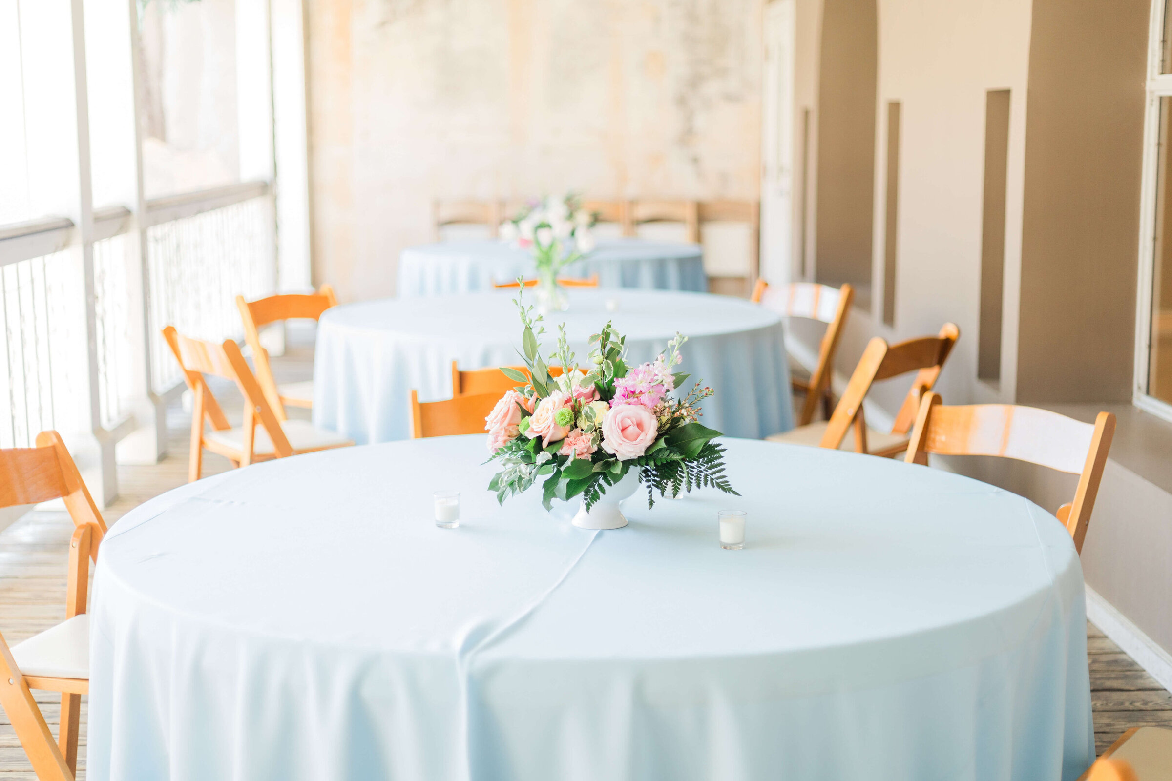 Round tables with light blue tablecloths and wooden chairs are set up outdoors. Each table has a floral centerpiece and place cards. Sunlight creates warm shadows on the scene.