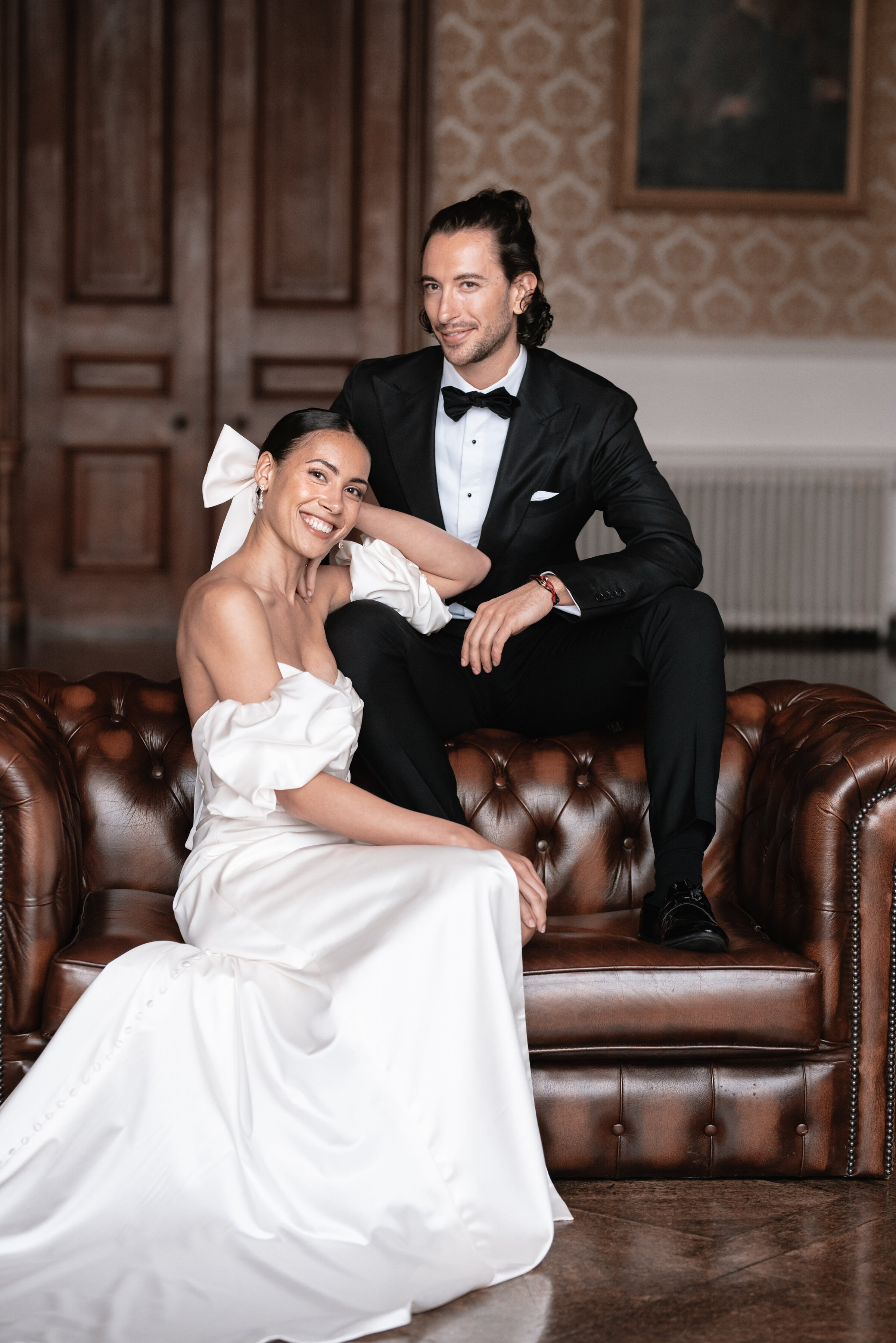 Bride and Groom in wedding attire seated on a leather sofa, smiling directly at the camera