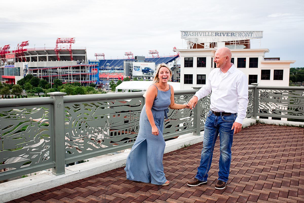 Girl wearing a blue dress and holding her fiances hand on the pedestrian bridge in Nashville with Nissan stadium in the background