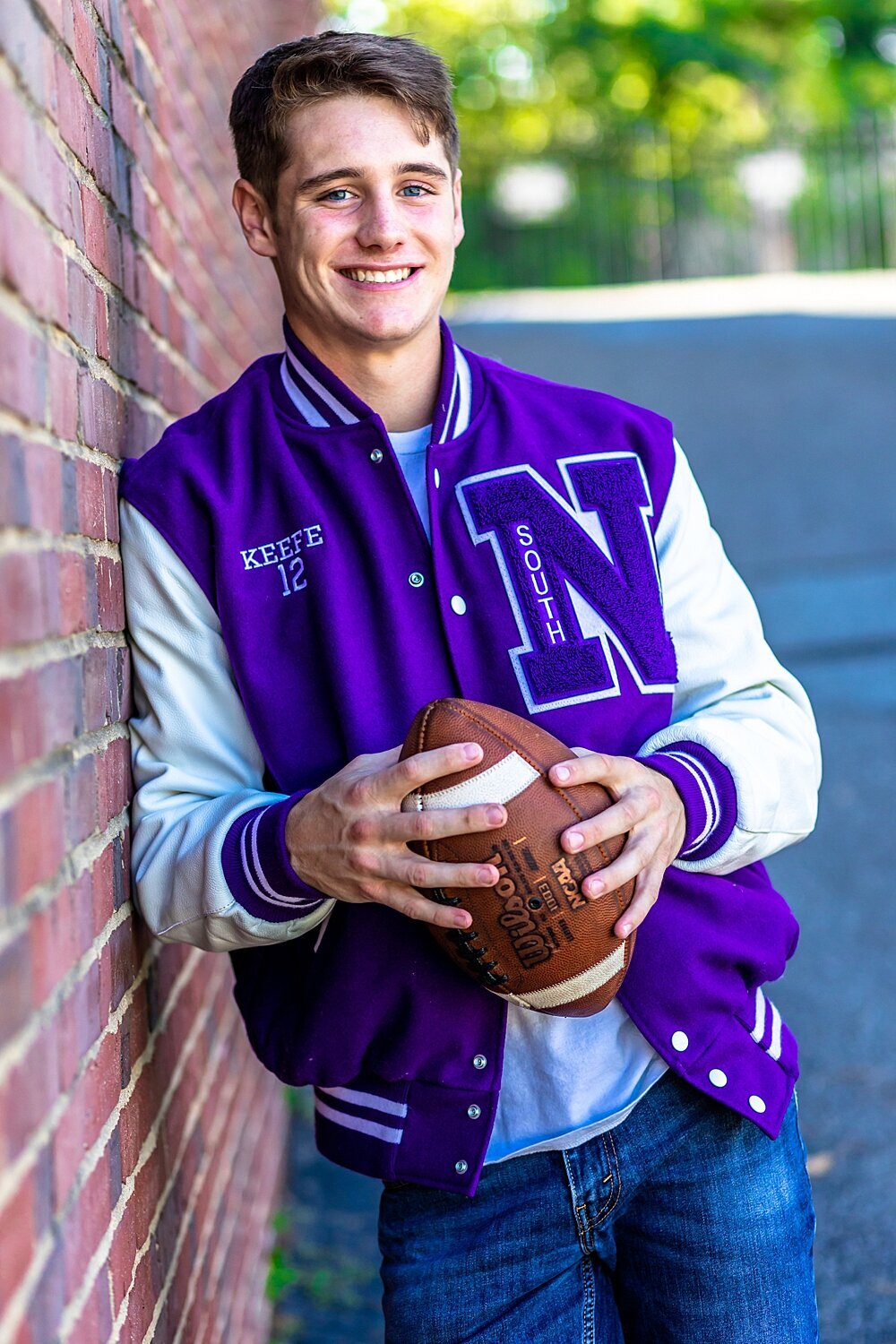 Senior boy leans against brick and holds football while wearing senior football jacket at his senior session in Nashua, New Hampshire