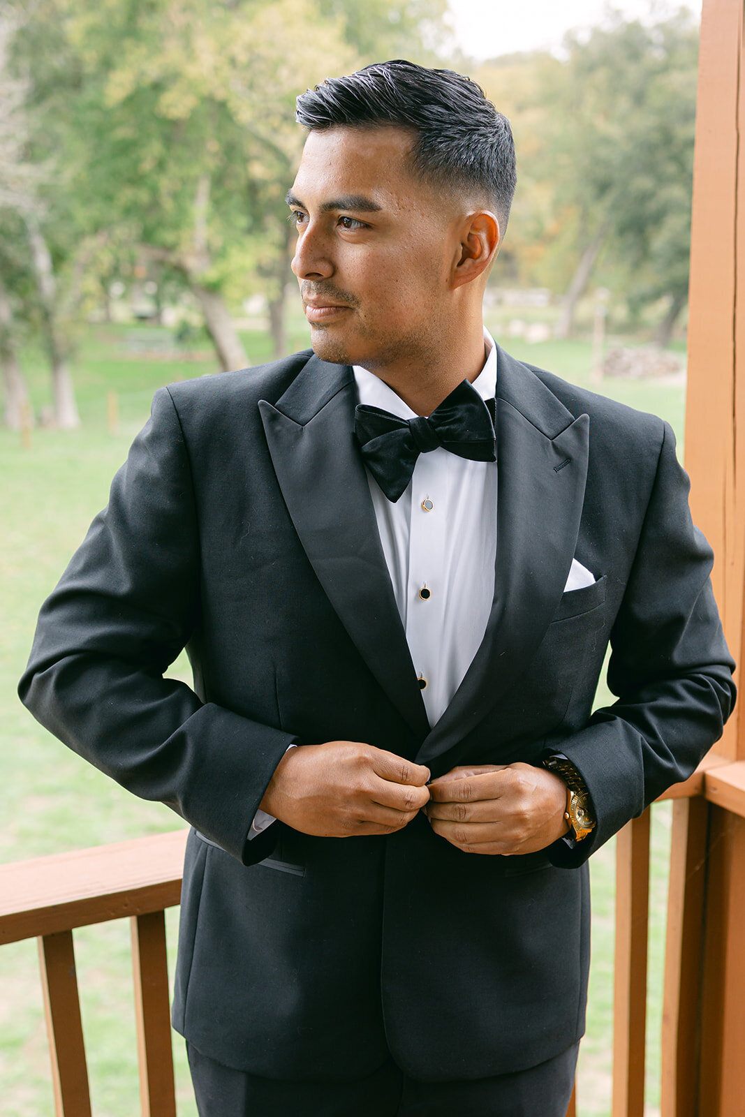 Groom in a sharp black tuxedo adjusting his jacket while standing on a wooden balcony at an upscale Austin wedding venue, preparing for the ceremony