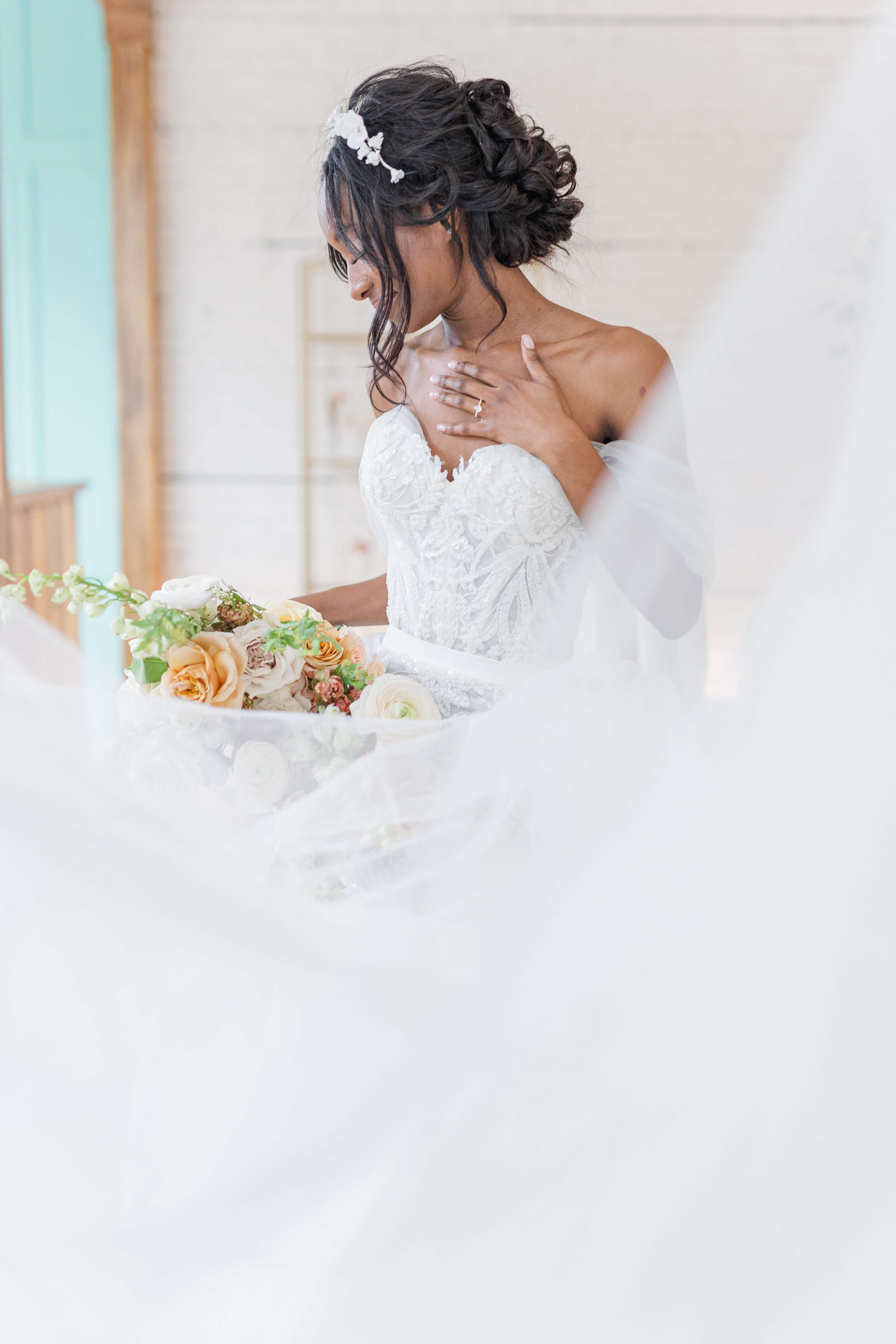 A bride has her dress blowing as the looks down with her hand on her chest. there is a light blue and white wall in back of her.