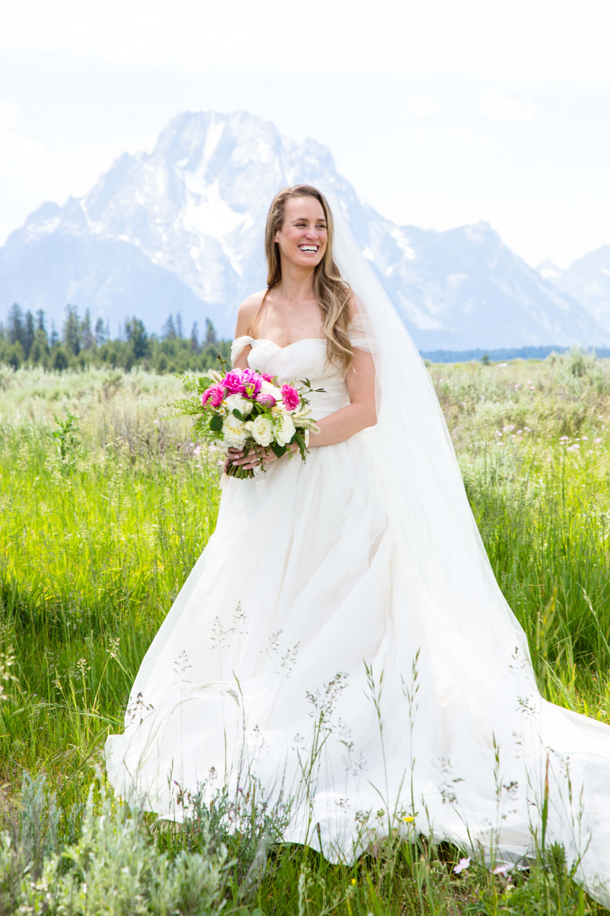 A bride stands in a lush meadow at Grand Teton National Park, holding a vibrant bouquet of pink and white flowers. She smiles against the backdrop of Mount Moran, wearing a flowing white gown with a long veil.