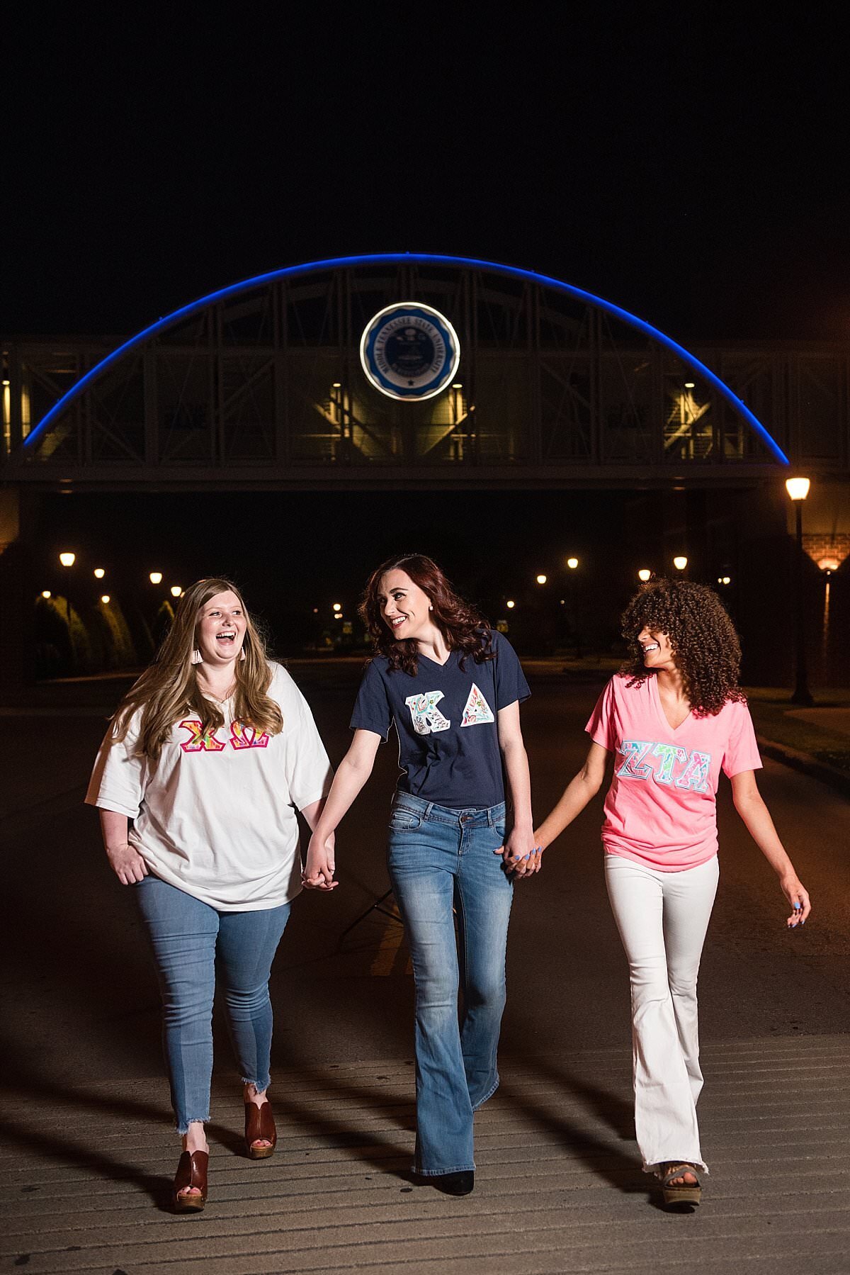 Chi Omega, Kappa Delta, Zeta Tau Alpha Seniors holding hands and walking underneath the bridge on the MTSU campus at night while it is lit blue