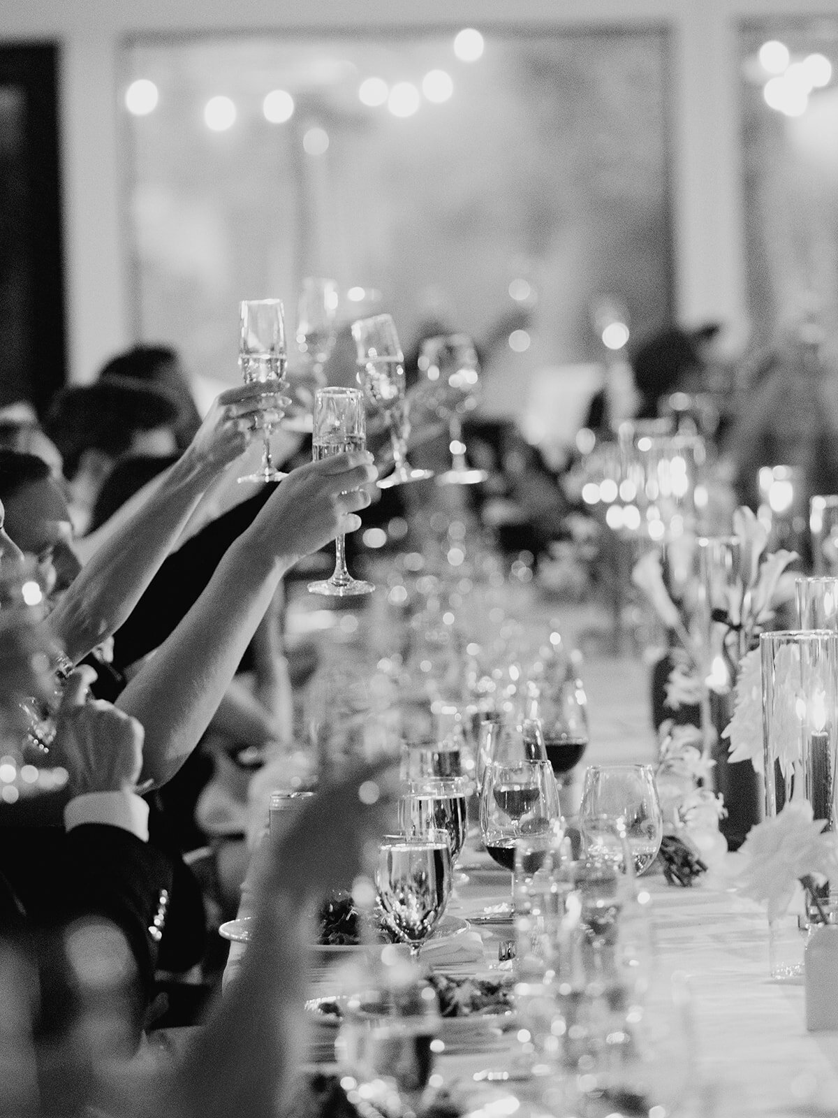 Black and white photo of wedding guests raising their glasses along a long candlelit table