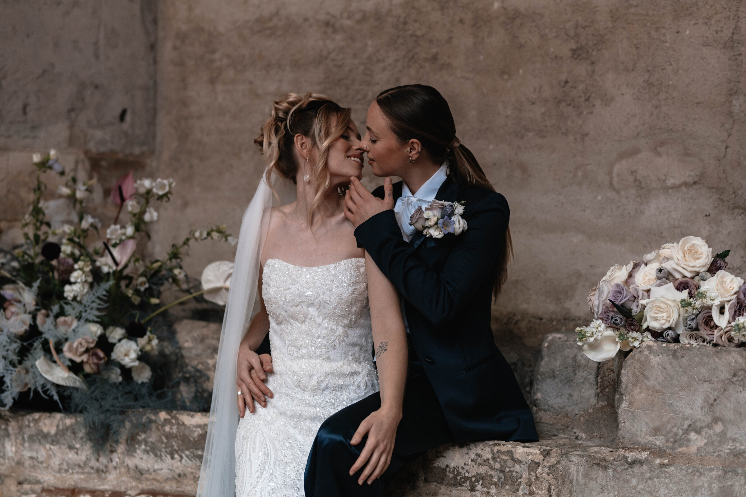 Two brides in wedding attire surrounded by flowers just about to kiss