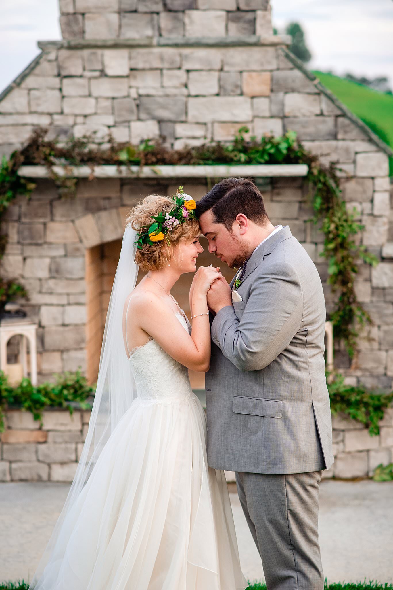 Newlyweds whispering to one another on the hillside of White Dove Barn in front of the outdoor fireplace, she is wearing a flower crown