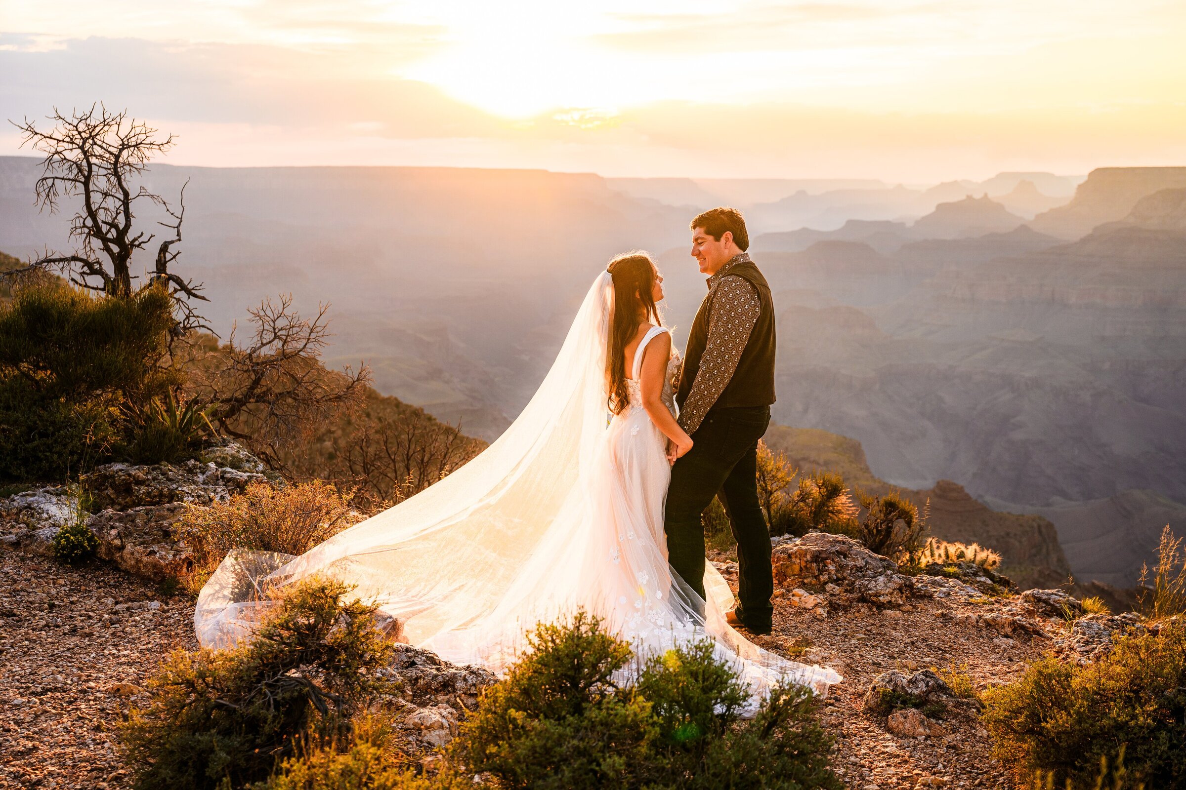 bride and groom standing on edge looking at each otherat the Grand Canyon with golden hour sunset behind them