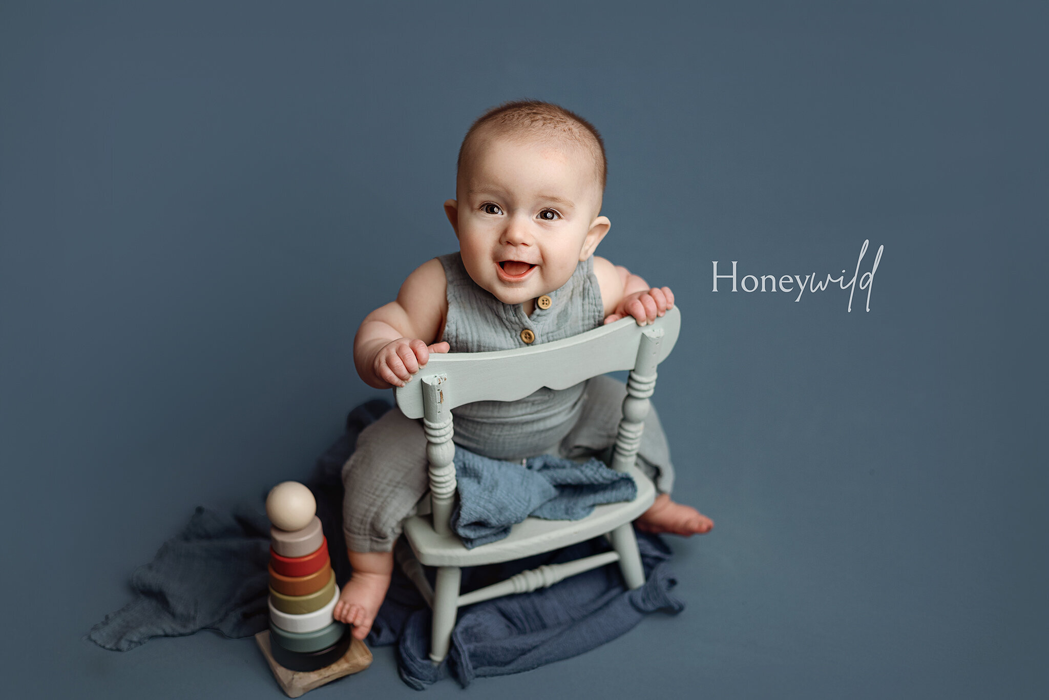 Grand Rapids newborn baby photography by Honeywild Photo: smiling six month old sitting on a mint chair backwards on a beautiful rich blue background with a wooden stacking toy near his feet