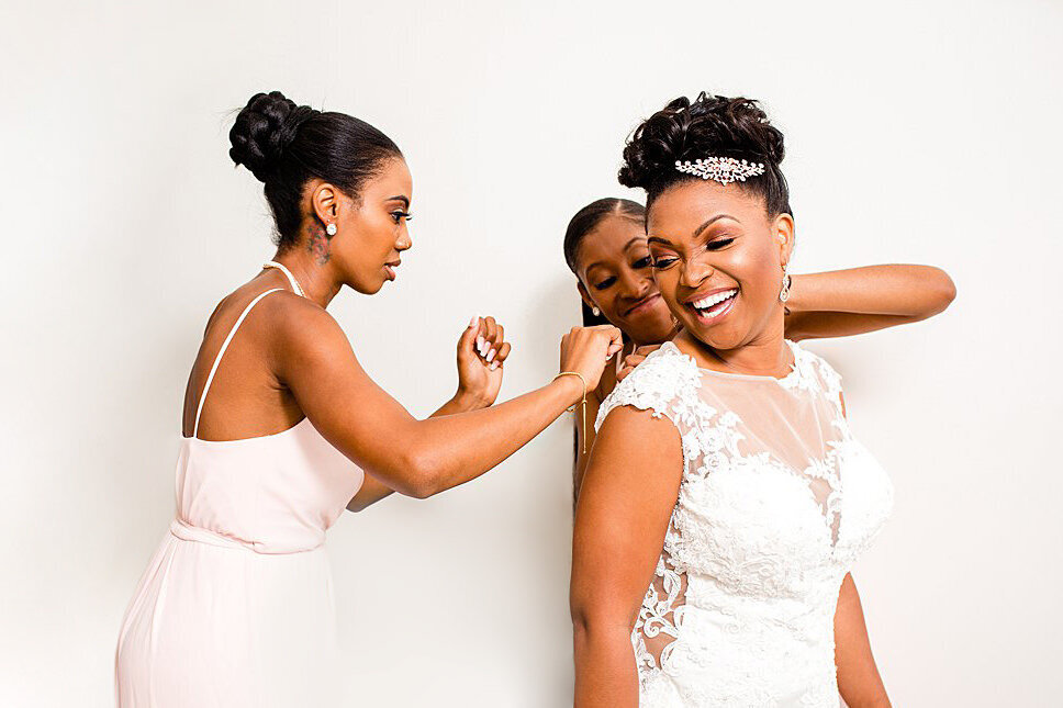 Bridesmaids laughing with bride while buttoning up her dress