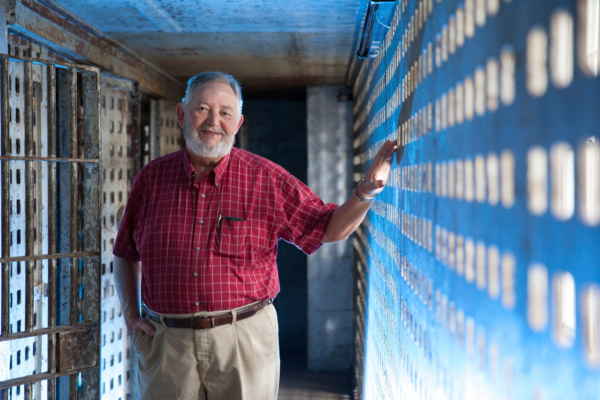 An older man with a beard, in a red plaid shirt and beige pants, stands in a narrow corridor with a metal bar wall. He smiles, resting one hand on the wall as soft lighting creates a grid of shadows—captured by a Jacksonville headshot photographer's keen eye.