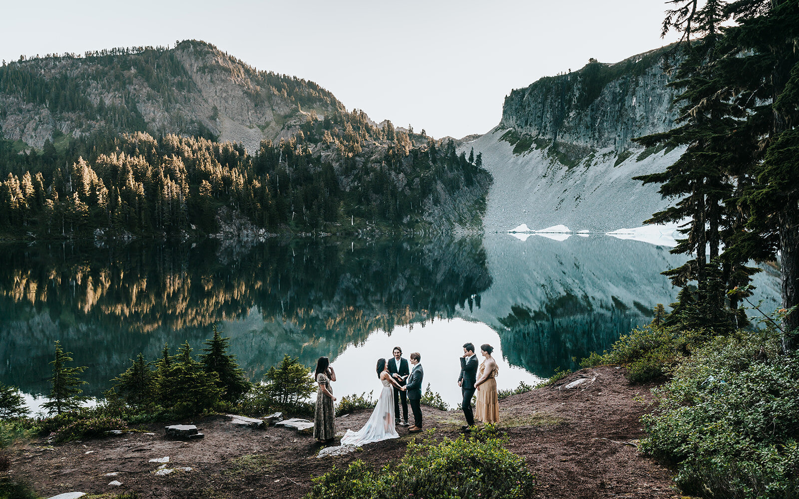 a bride, groom and their officiant are surrounded by three guests as they exchange vows on the shore of an alpine lake. mountains rise above them during their ceremony. Their adventure elopement photographers shoot from a higher vantage point showing the striking landscape. The mountains are reflected in the still waters and our couple, with their guests are centered perfectly in the negative space of the mountains 