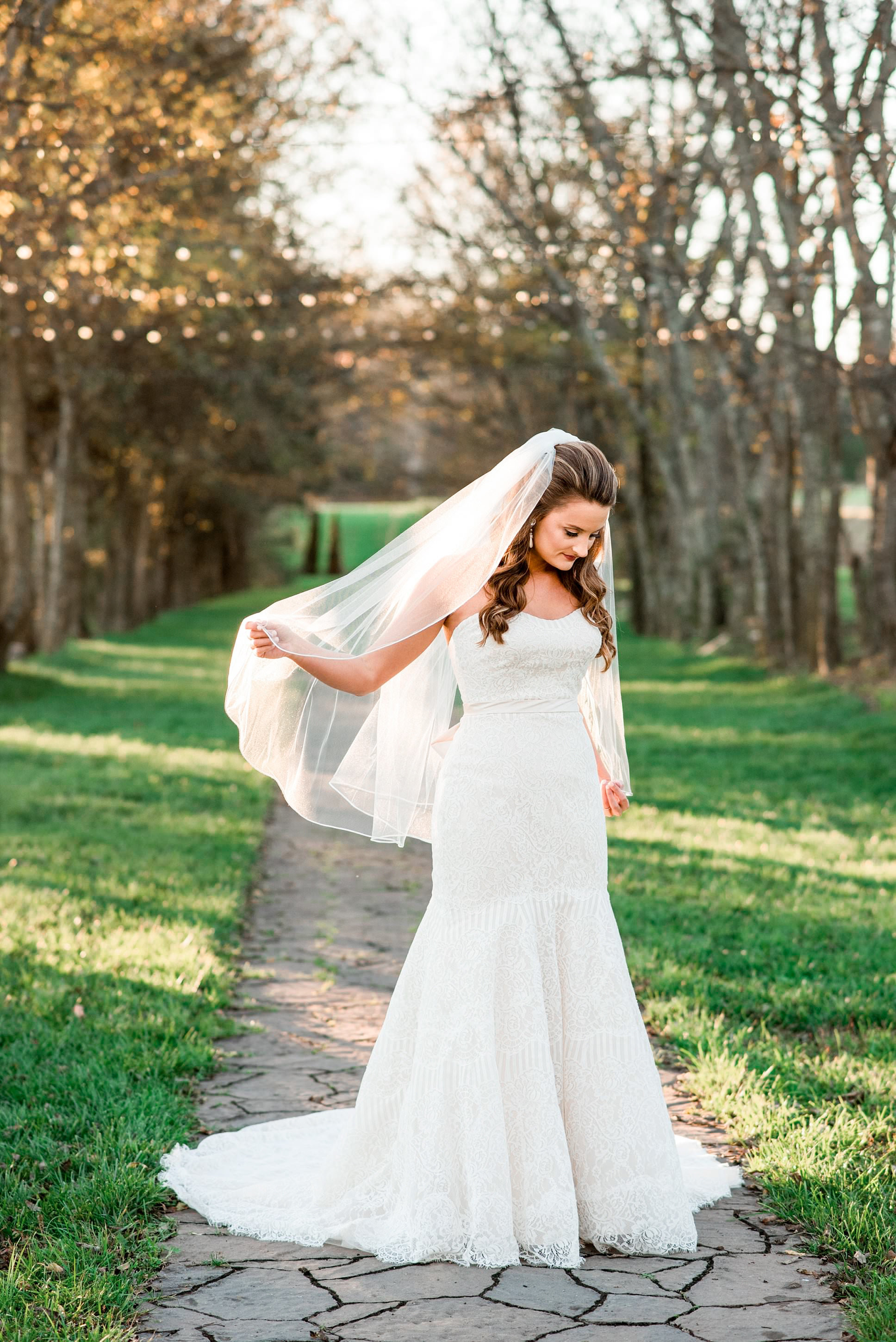 Bride standing on the sidewalk at Lovers Lane playing with her veil, bistro lights hanging overhead