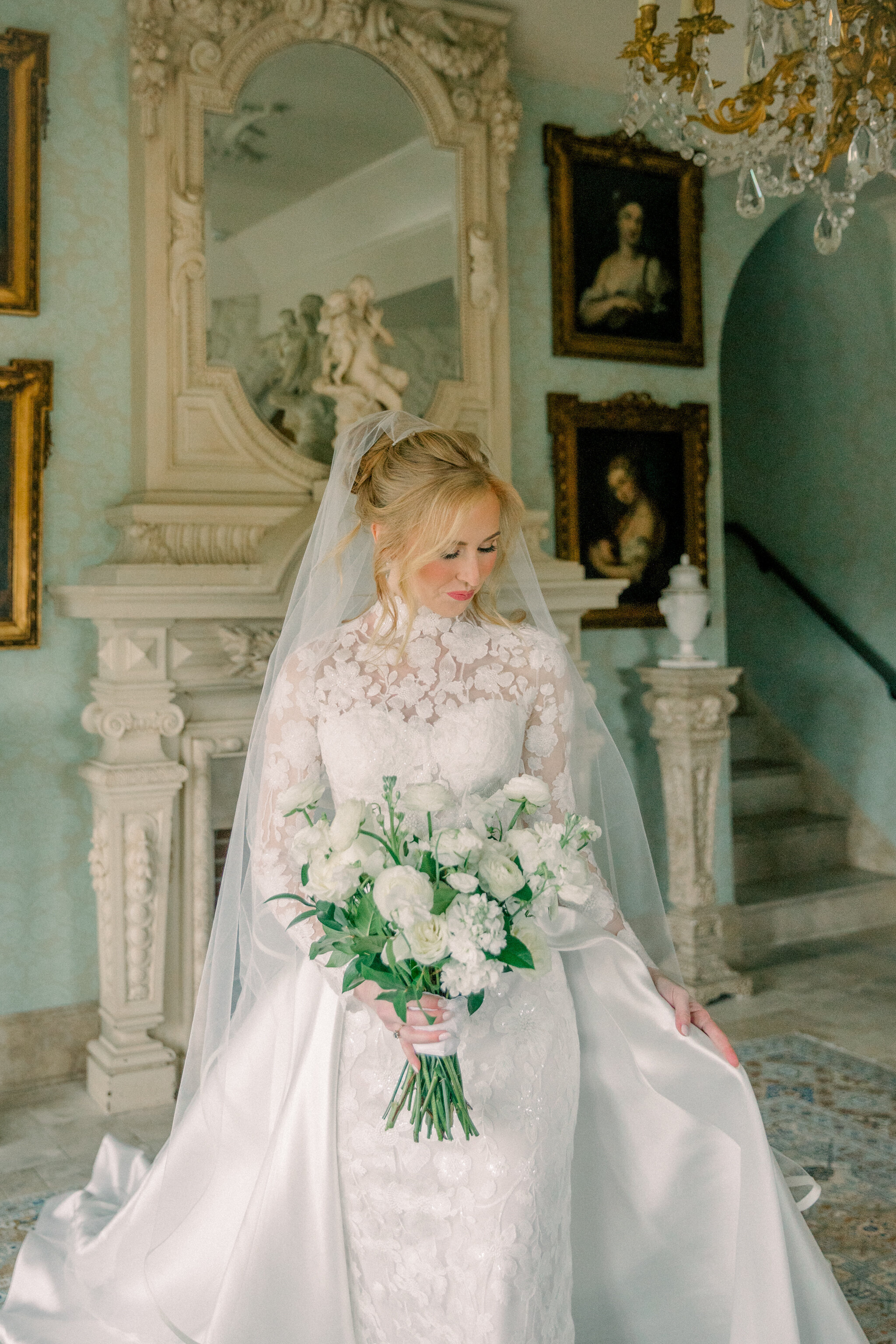 A bride in an elegant high-neck lace gown with a flowing satin train stands in the opulent interior of Dover Hall in Richmond, Virginia. She holds a lush white bouquet, framed by antique paintings and ornate architectural details. Captured by Caitlin Wilcox Photography, a Dover Hall wedding photographer specializing in romantic film and digital imagery.
