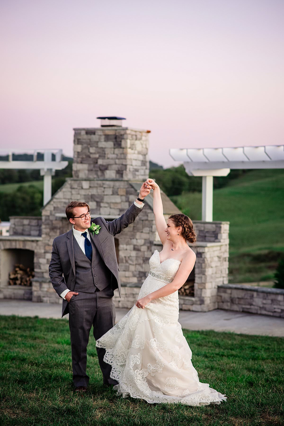 Groom holding his wife's hand as she twirls in her wedding dress in front of the outdoor stone fireplace at White Dove Barn during sunset