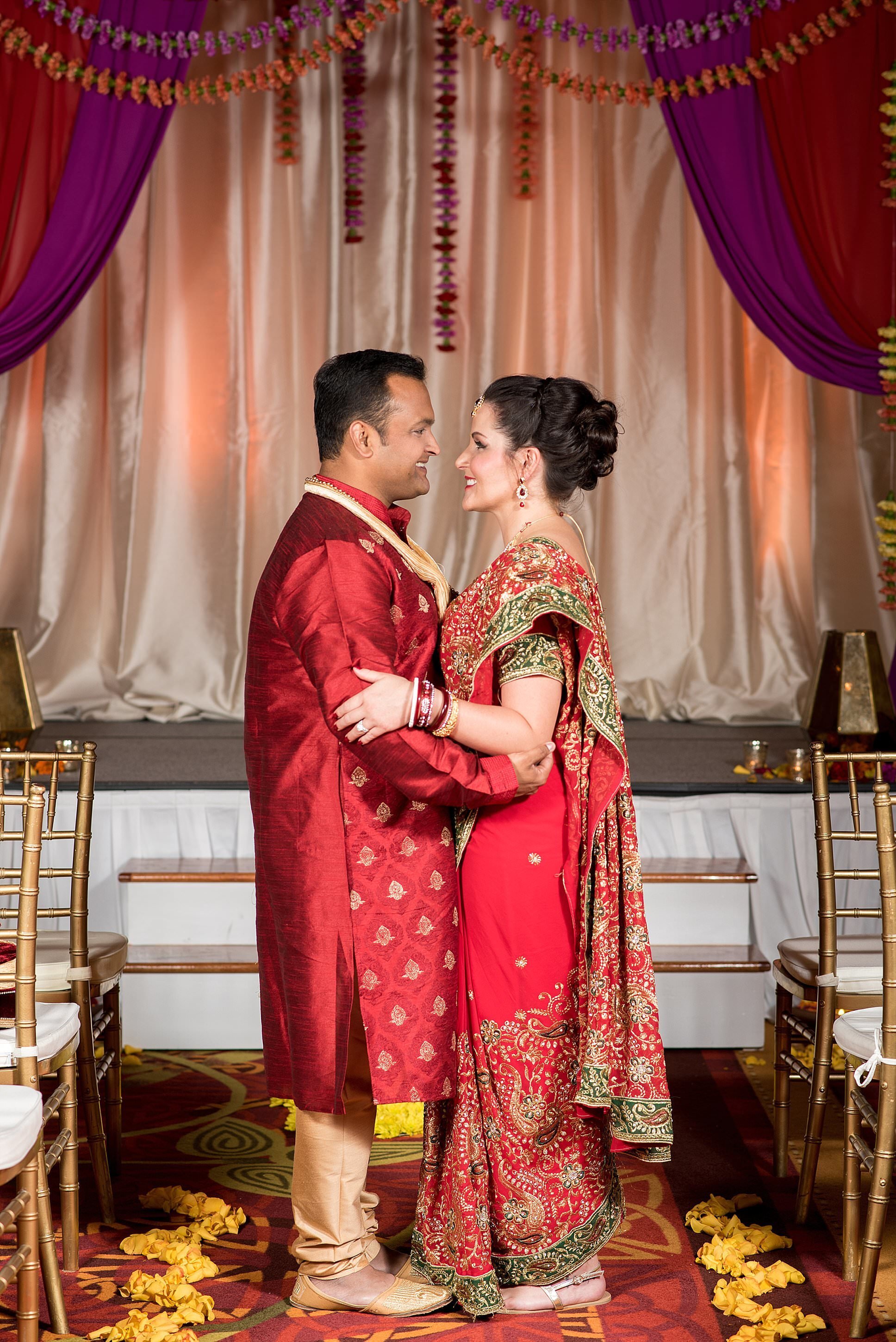 Couple facing one another under fuchsia and red mandap wearing traditional Indian wedding attire