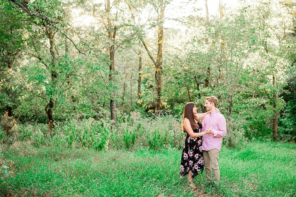 Couple dancing together in a field with trees surrounding them. She is wearing a long black dress with a pink floral pattern, he has on a pink dress shirt