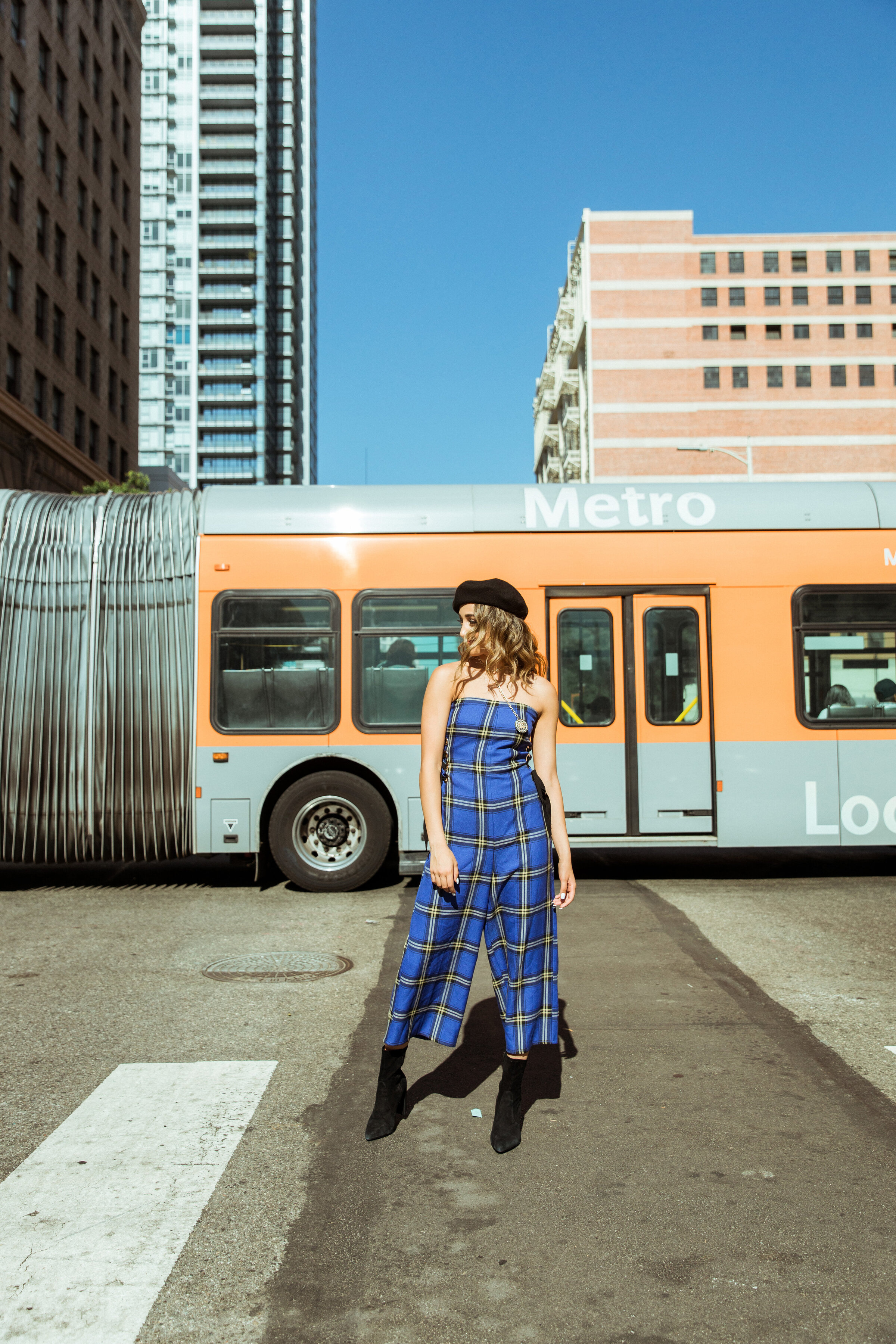 Gritty street-style image of a young woman standing confidently in front of a Metro Los Angeles bus, wearing a bold blue plaid jumpsuit and black beret, channeling edgy downtown fashion energy.
