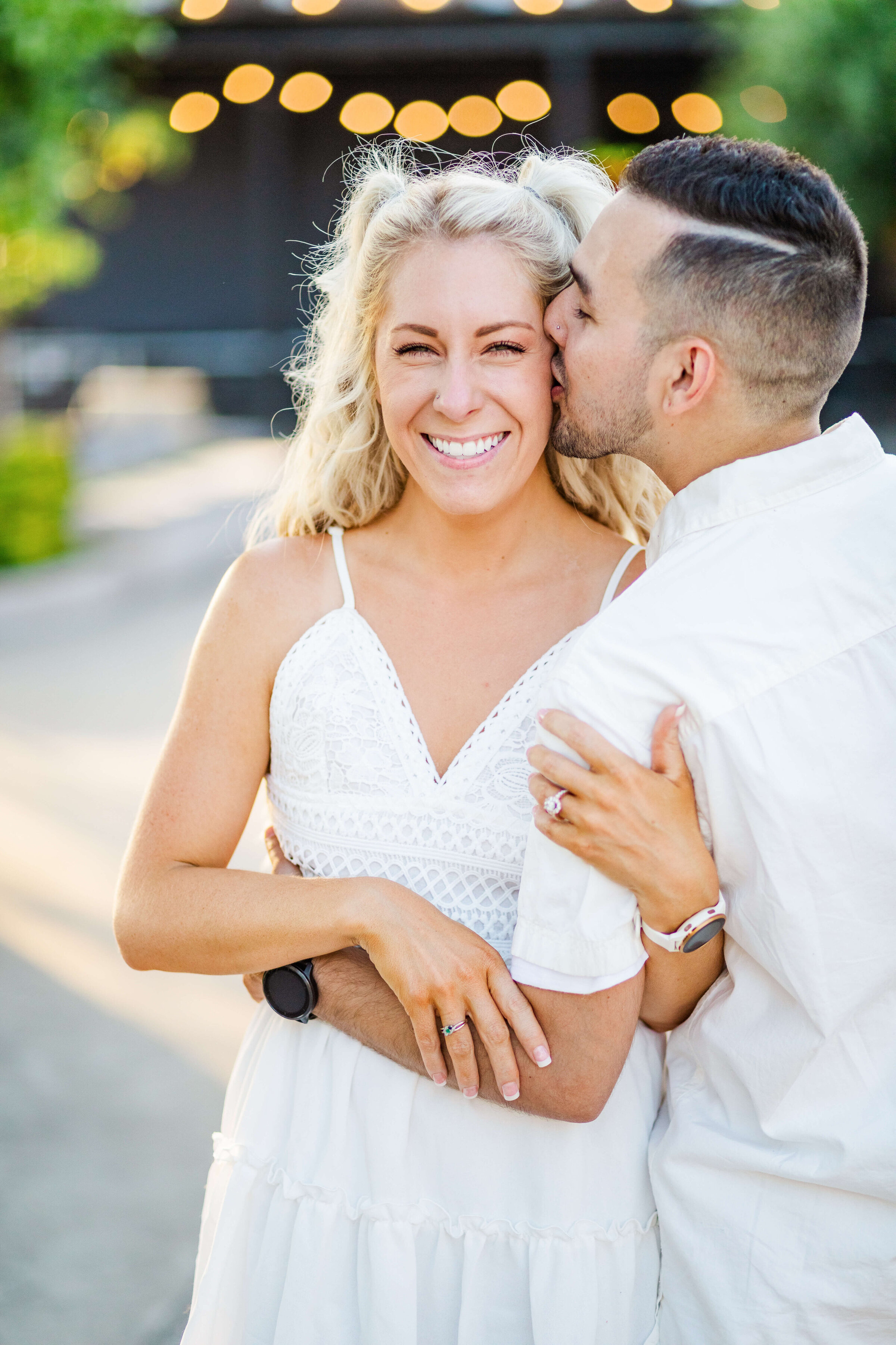 A woman is smiling as her fiance hugs and kisses her on the cheek. They are both wearing white.