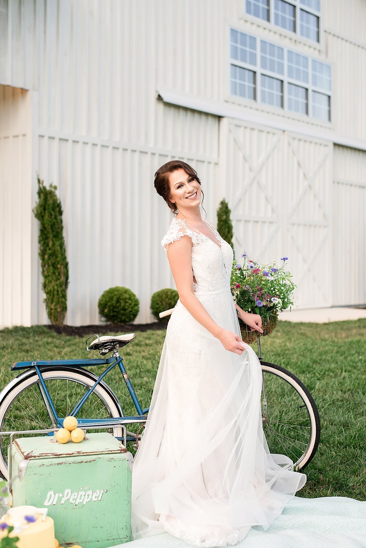 Bride standing beside vintage bicycle with vintage cooler outside of the White Dove Barn
