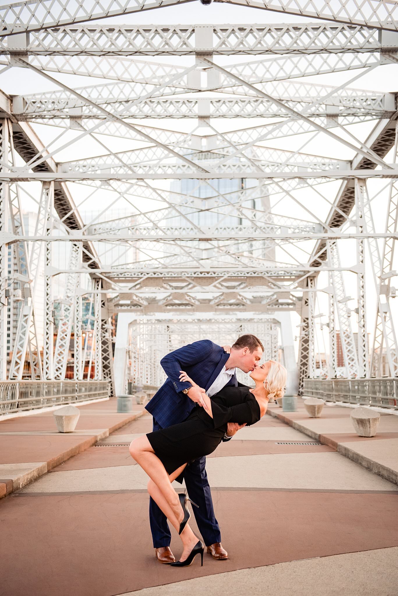 Man dipping his fiance while standing atop the Nashville pedestrian bridge, he's in a blue suit and she is wearing an elegant black dress and heels