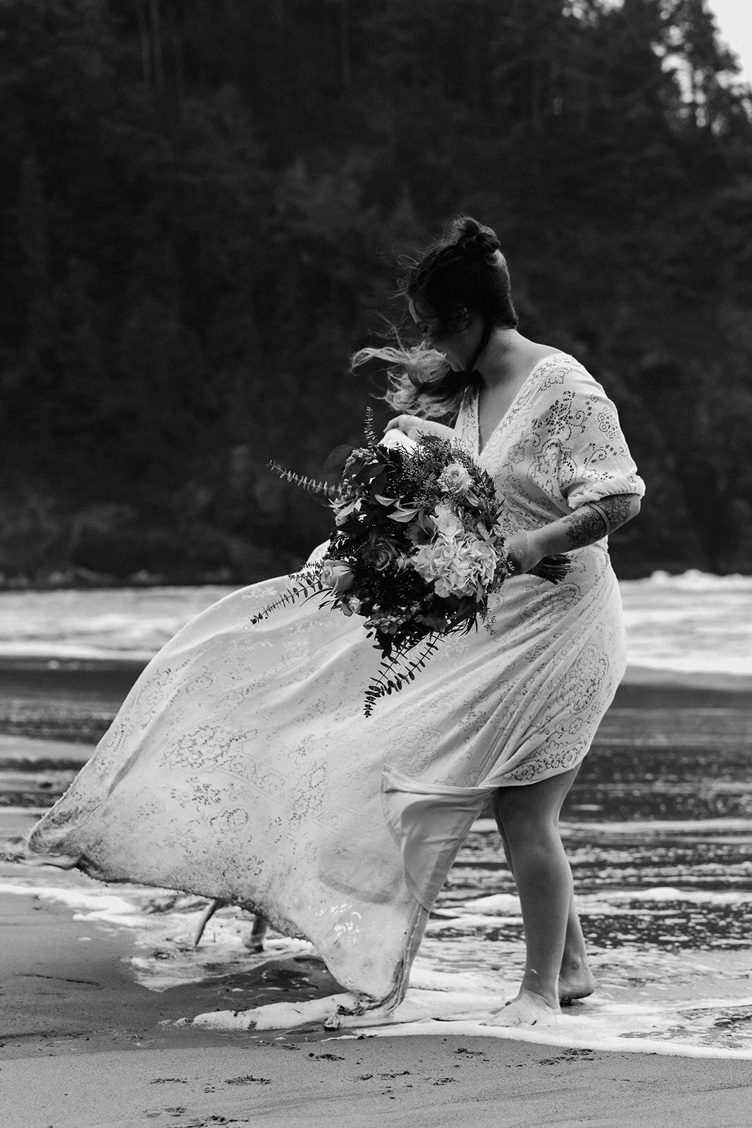 A black and white image of a bride walking out of the ocean. her dress and hair billow in the wind showing off the environment from this moody adventure elopement. 