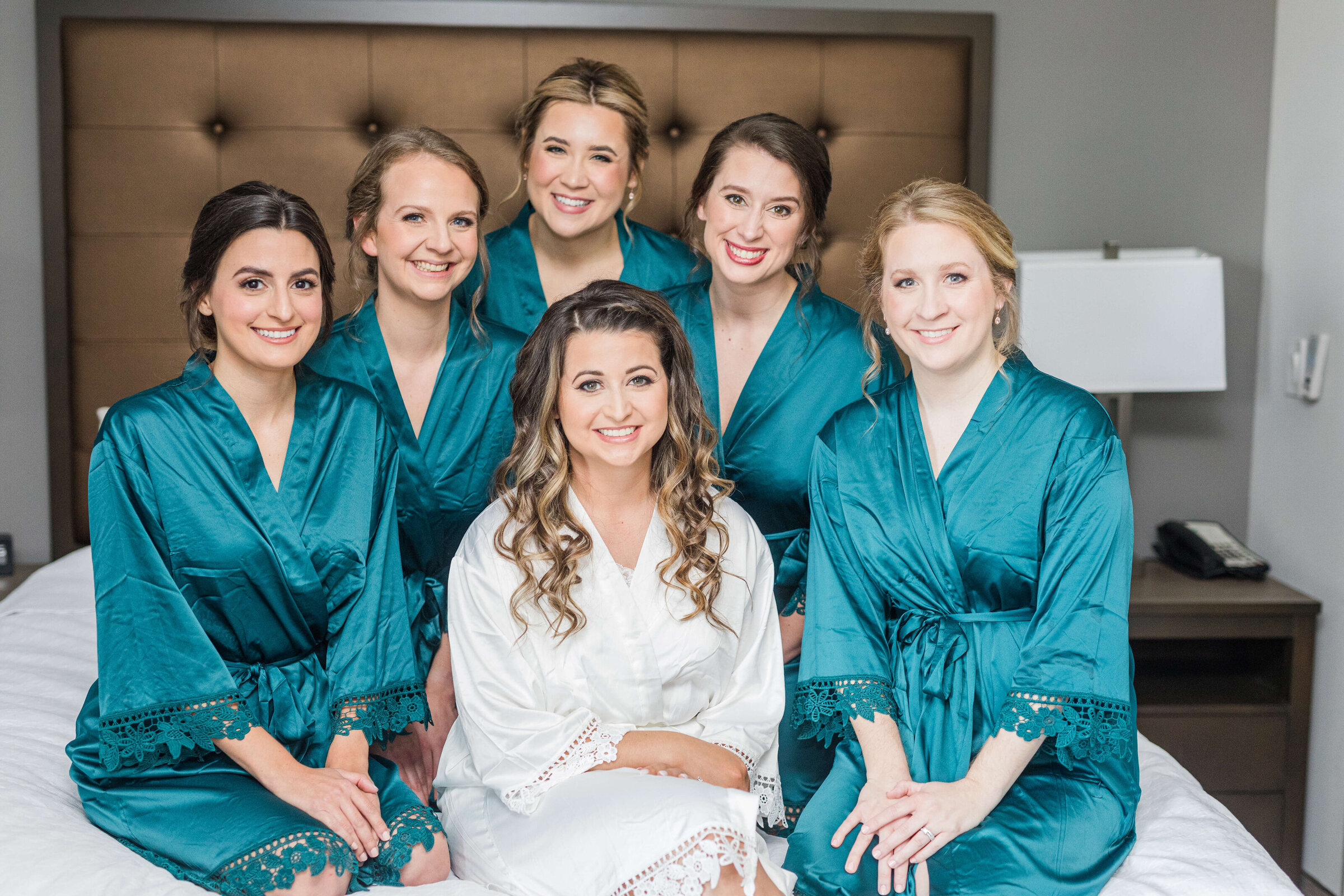 A bride in her white robe and bridesmaids in teal robes sit on a hotel bed smiling at the camera.