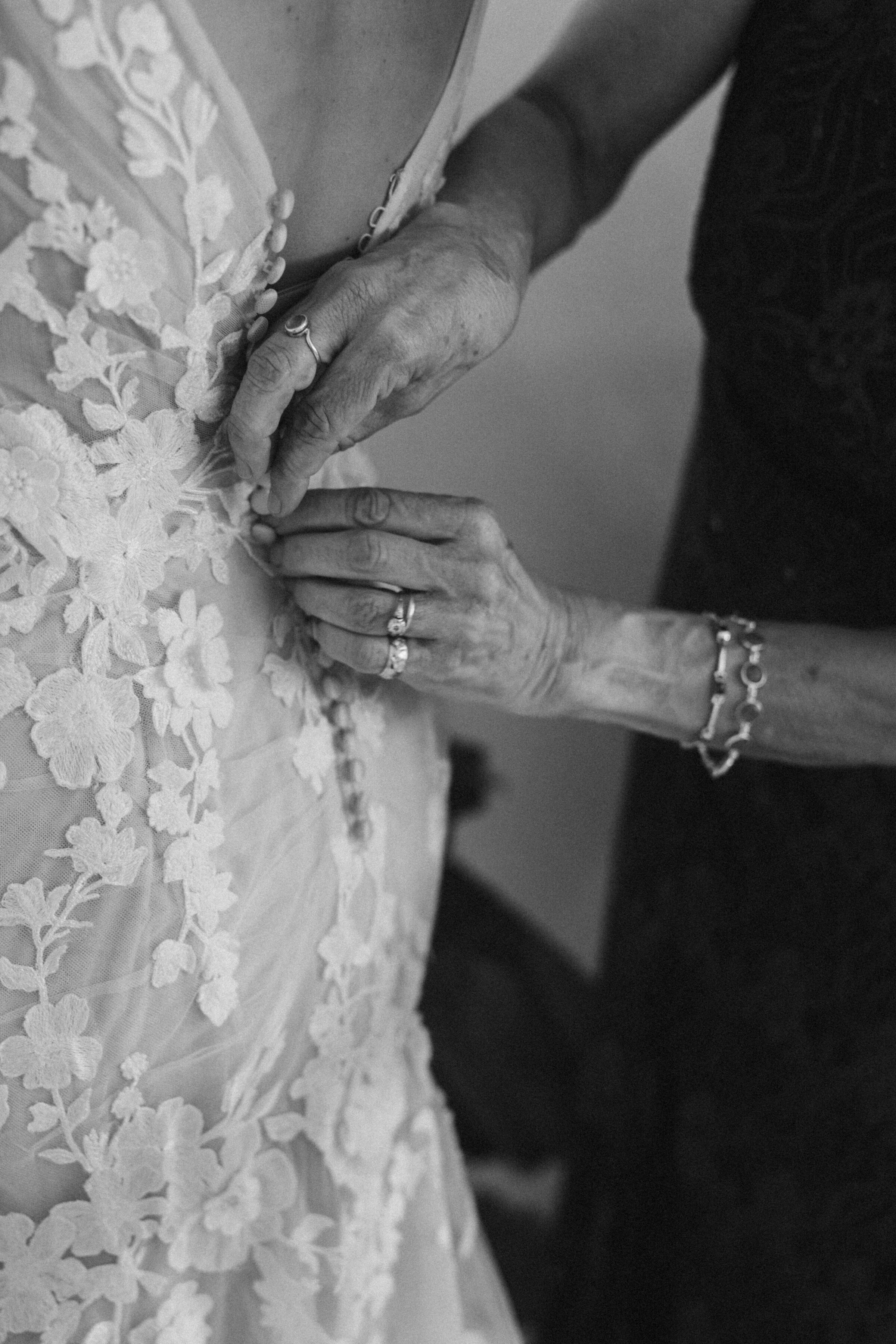 Close-up of an elder woman’s hands buttoning the back of a bride’s lace wedding dress, capturing a tender getting-ready moment in black and white.