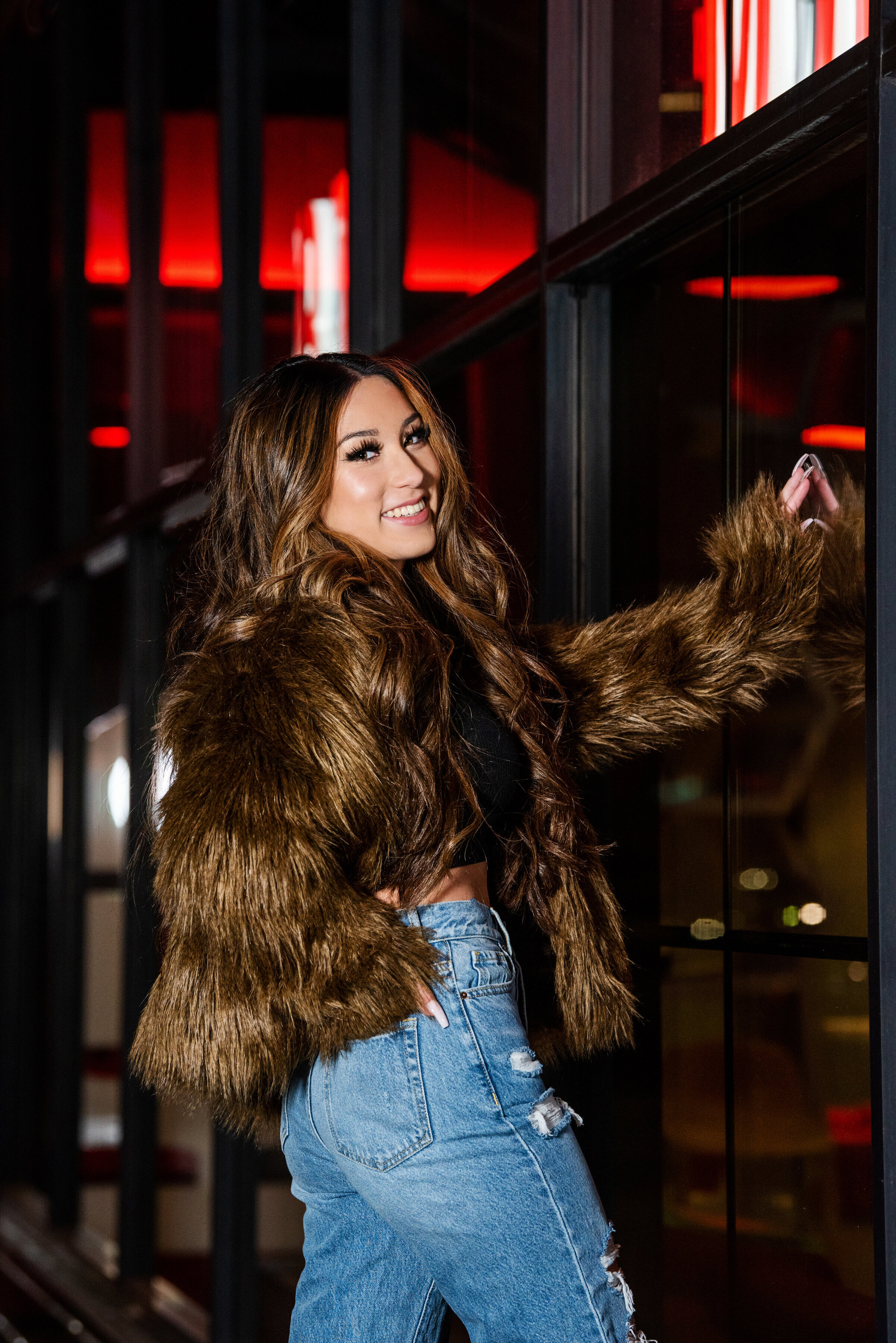high school senior photo  of girl in brown fur jacket leaning against glass at night in downtown minneapolis