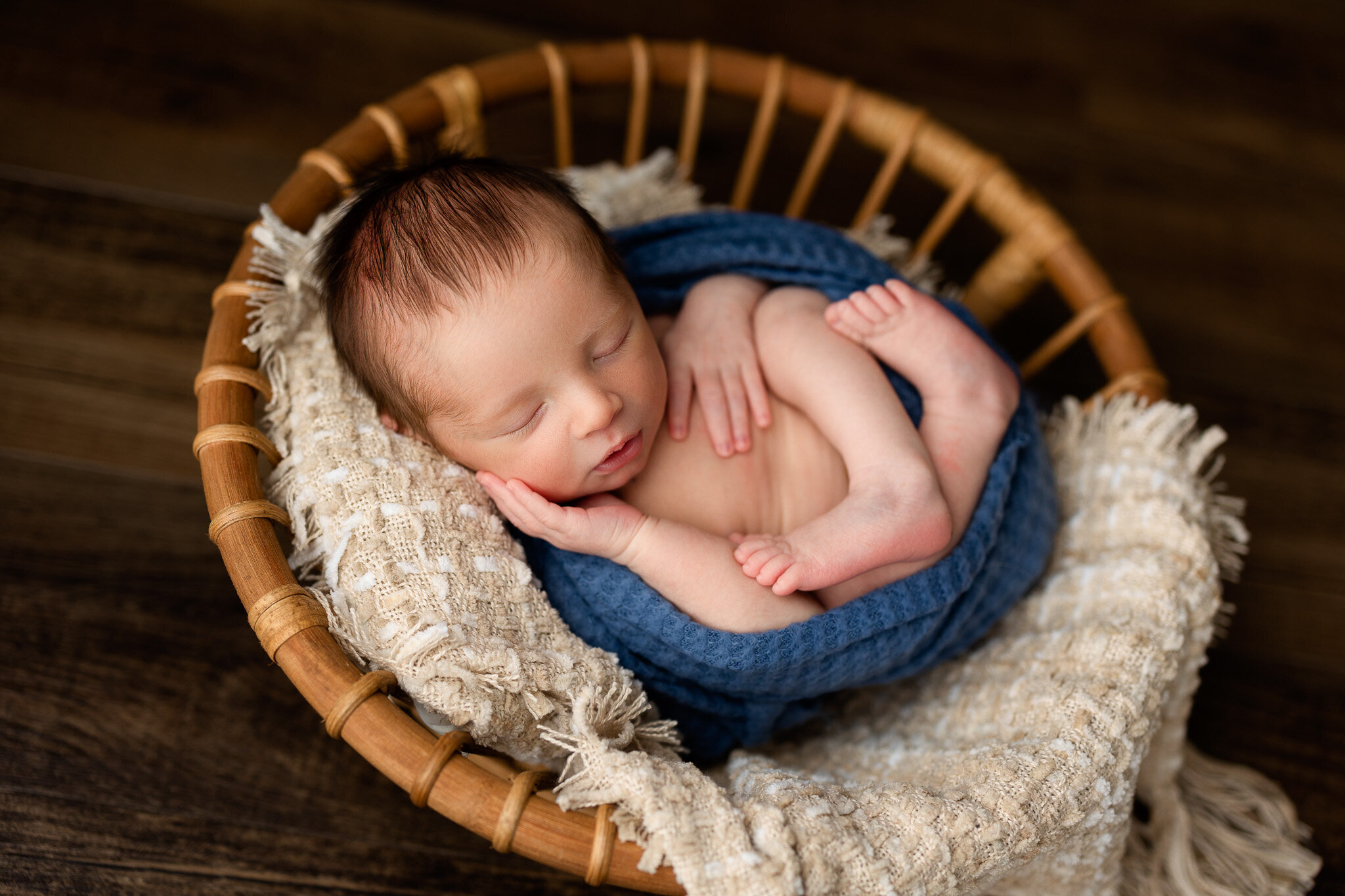 Newborn boy wrapped in blue posed in a wooden bowl