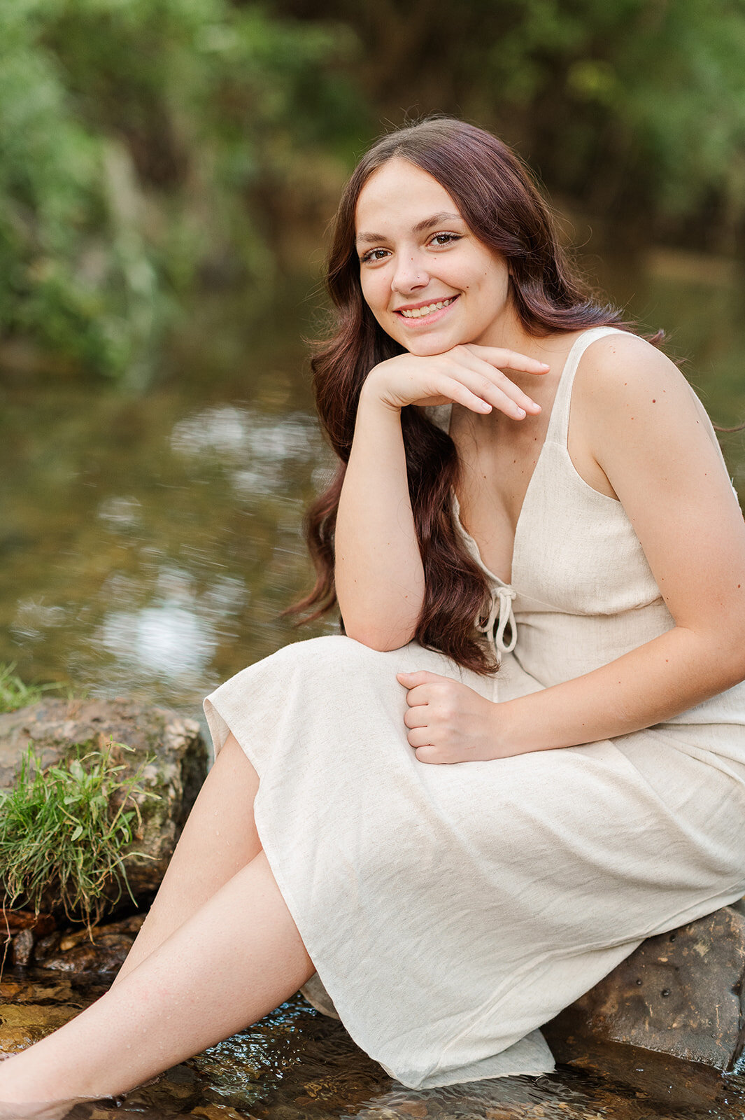 Senior girl sitting in a shallow creek wearing a light linen dress, smiling softly in natural light near York, PA

