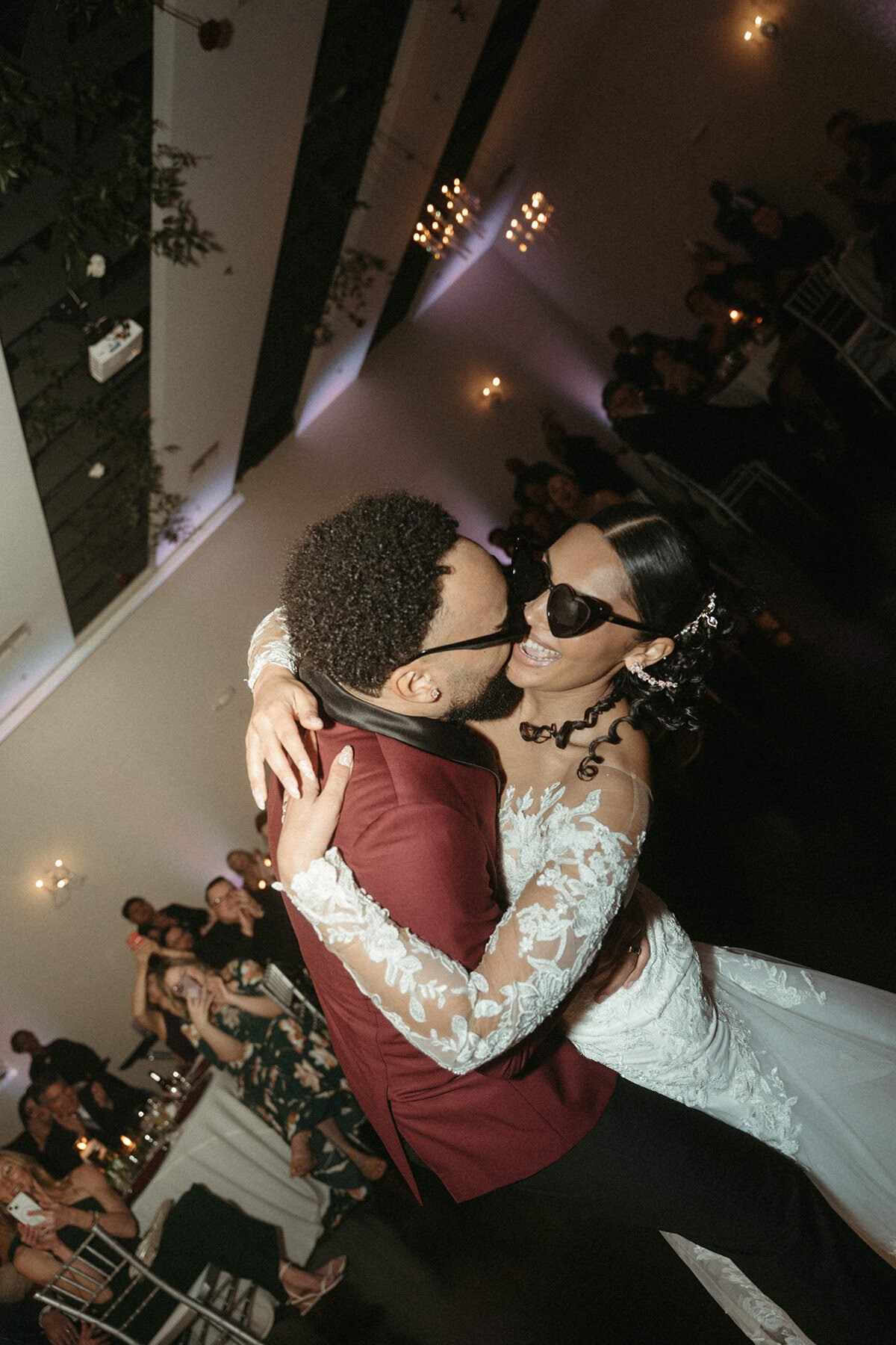 A bride in sunglasses and a lace wedding gown dances joyfully with her groom in a maroon suit at SoHo63, a Chandler wedding and event venue. The couple laughs together under twinkling lights, capturing the fun and modern vibe of weddings hosted at SoHo63.