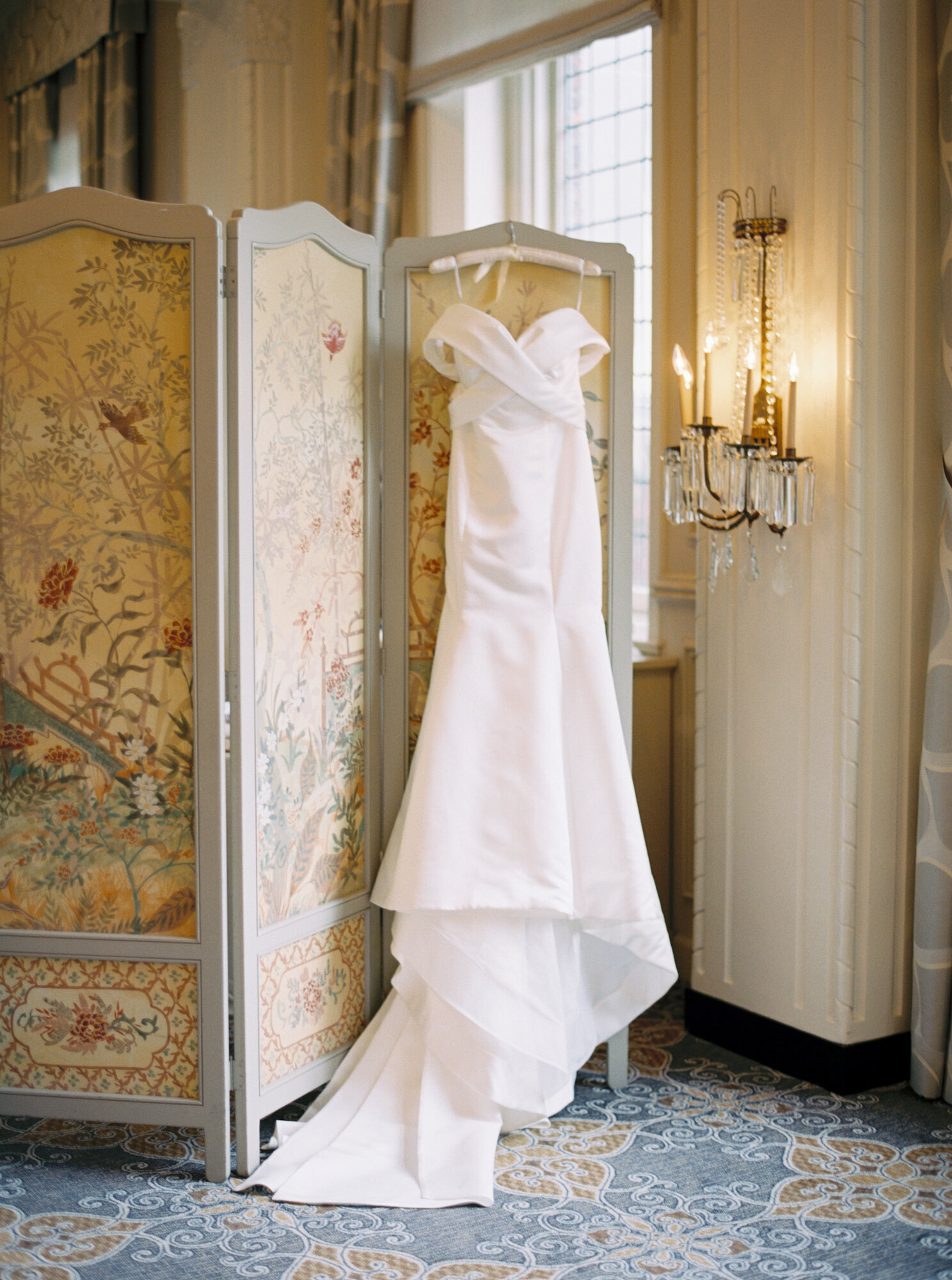 off the shoulder bridal gown hangs on a watercolor according wall in a historic ballroom