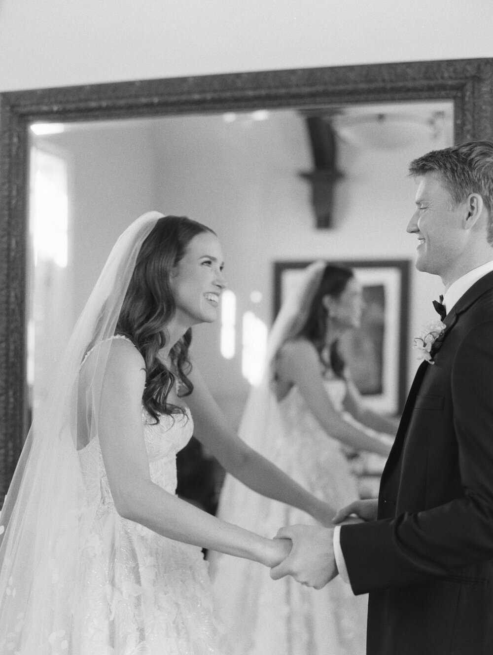 black and white film photo of bride and groom holding hands and smiling at each other candidly