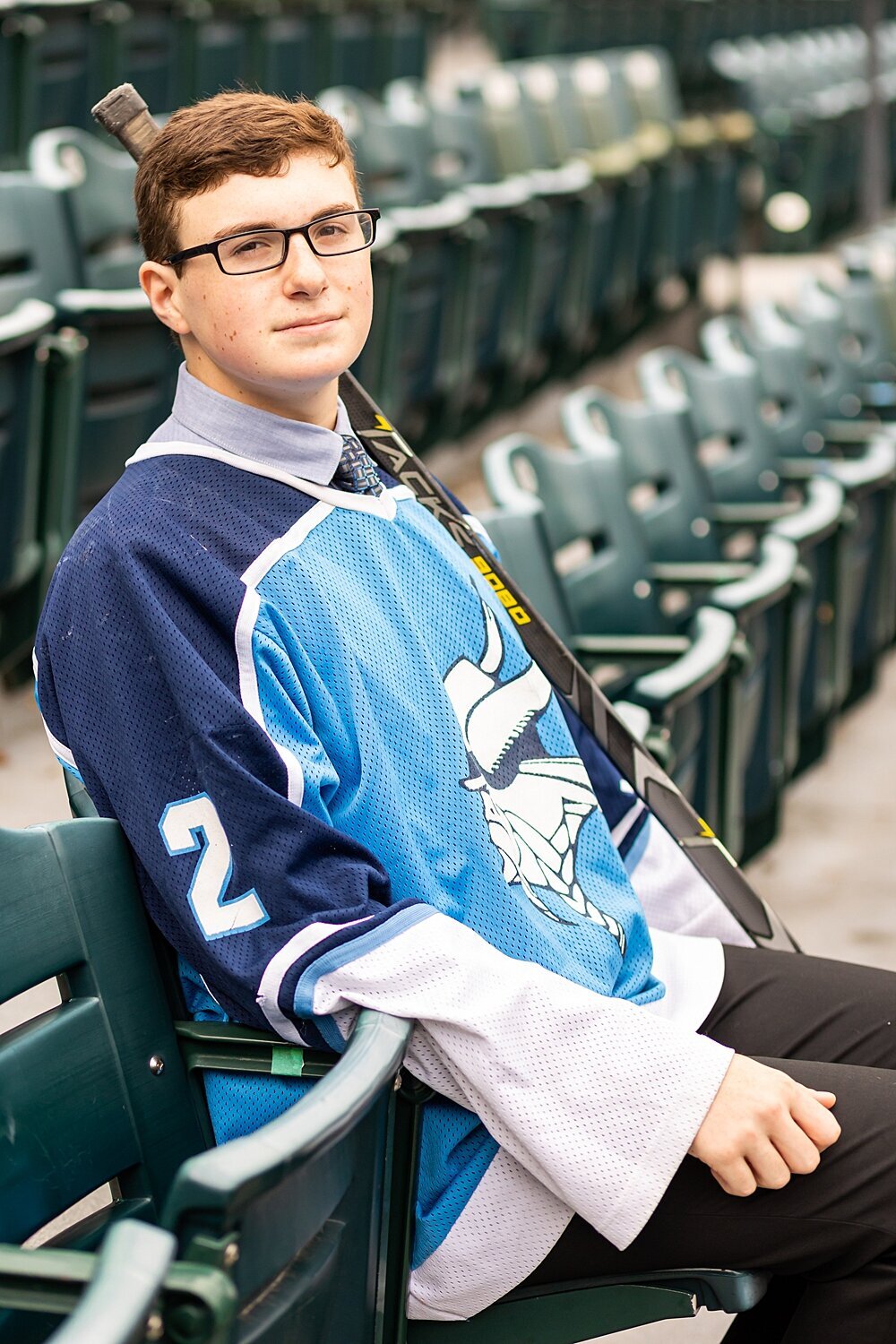 Senior boy sits in stadium seat with hockey stick and jersey during summer senior session in Nashua, New Hampshire