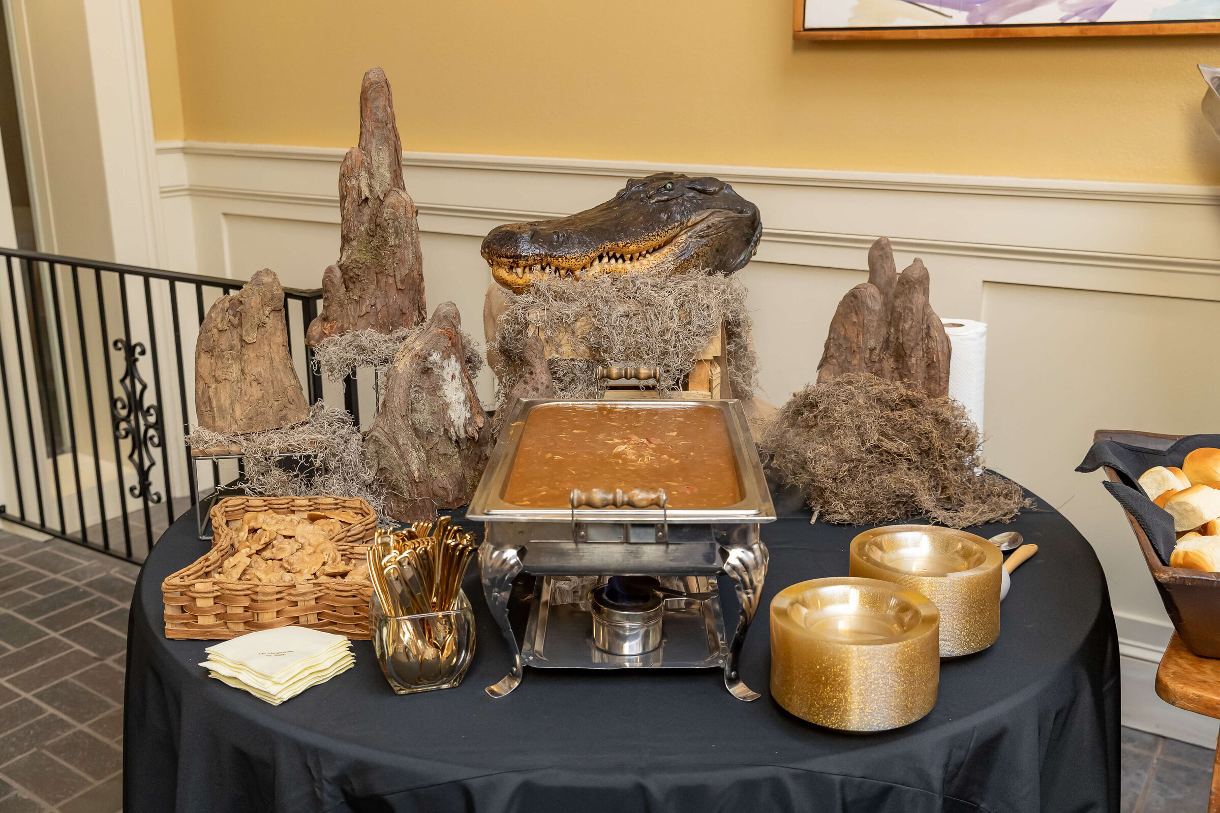 A buffet table decorated with a faux alligator head and rocks, featuring a tray of soup or stew, animal-shaped crackers, gold utensils, two gold bowls, and a basket of bread rolls.