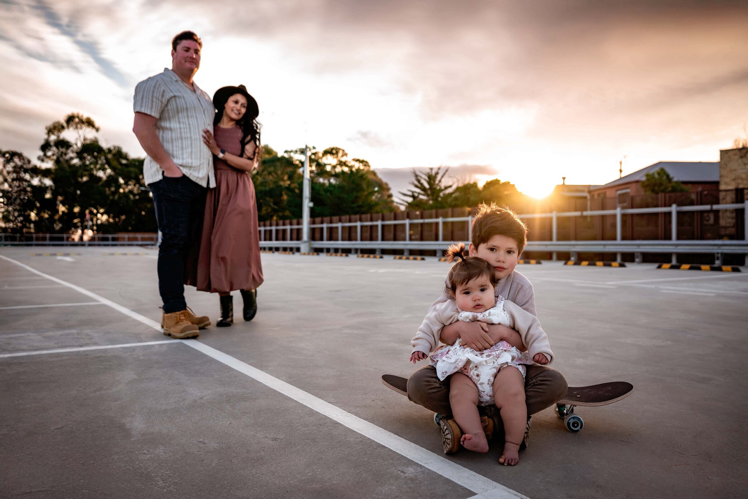 Rooftop family portrait taken at dusk with children seated on a skateboard and the parents looking on by Melbourne Family Photographer Kate Cashin