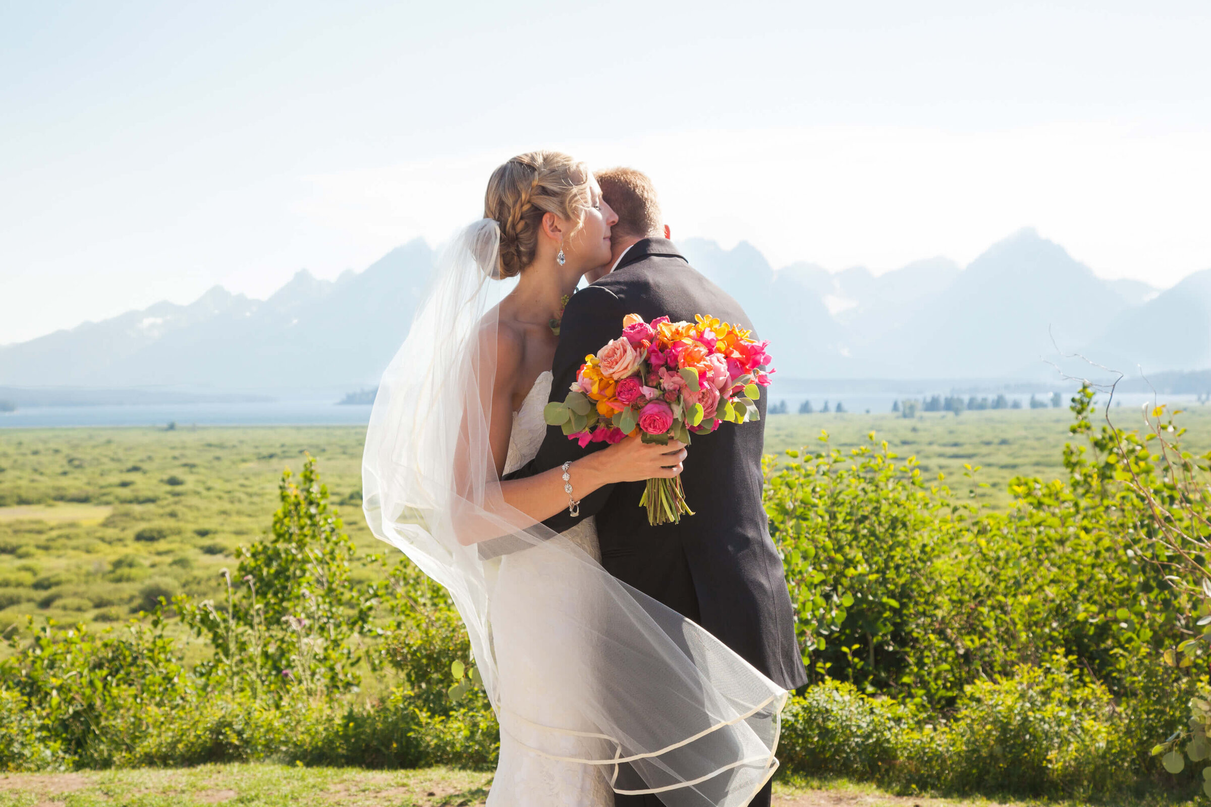 A bride in a lace gown hugs her groom in a black suit at Jackson Lake Lodge overlooking the Grand Tetons. The sunset casts a golden glow over the valley, creating a romantic Jackson Hole wedding scene.