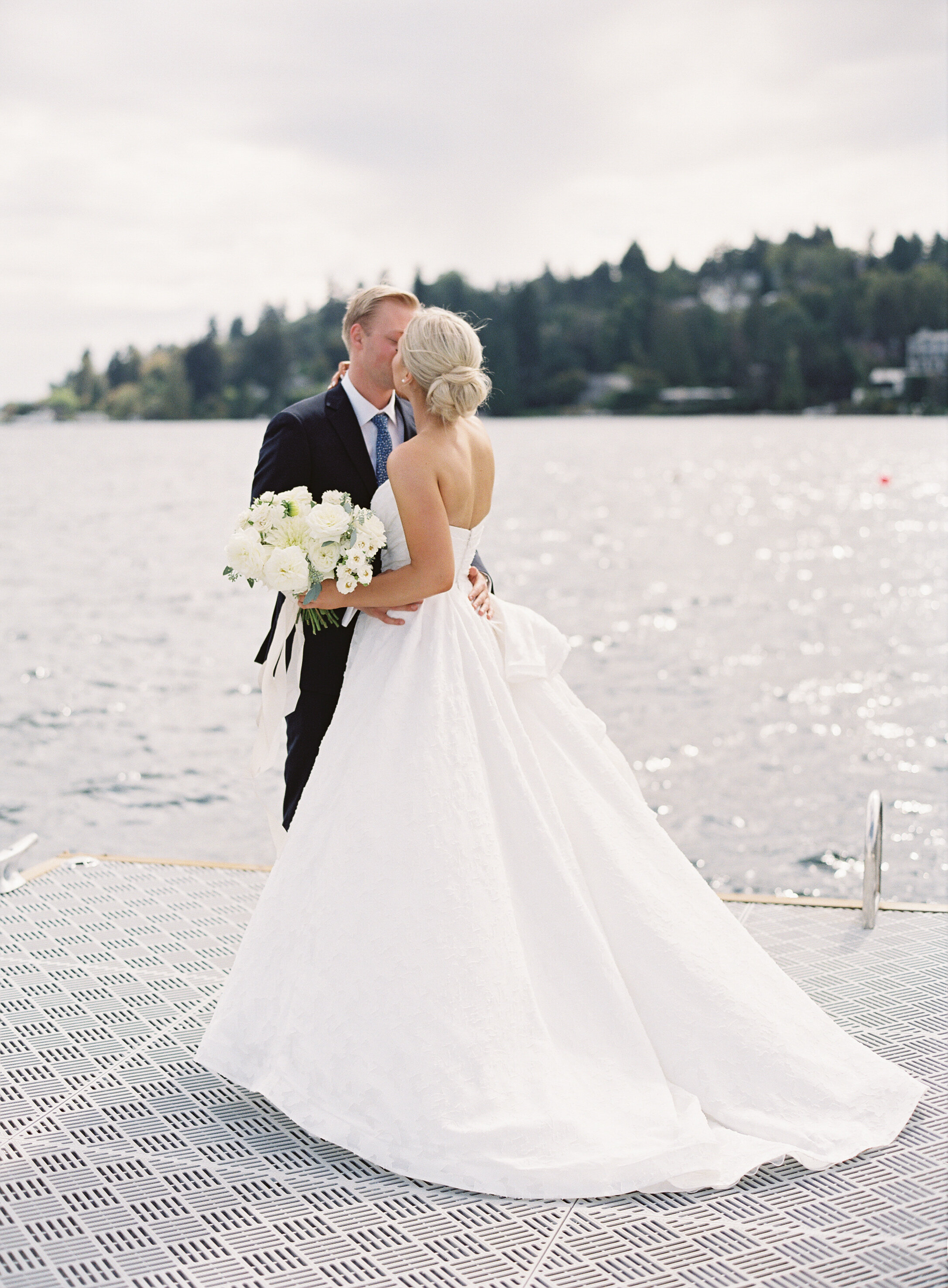 bride holds white rose boquet and kisses groom with back of dress showing on dock on lake