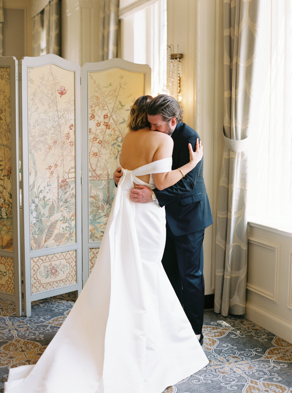 bride and groom hug at first look near large window in historic venue