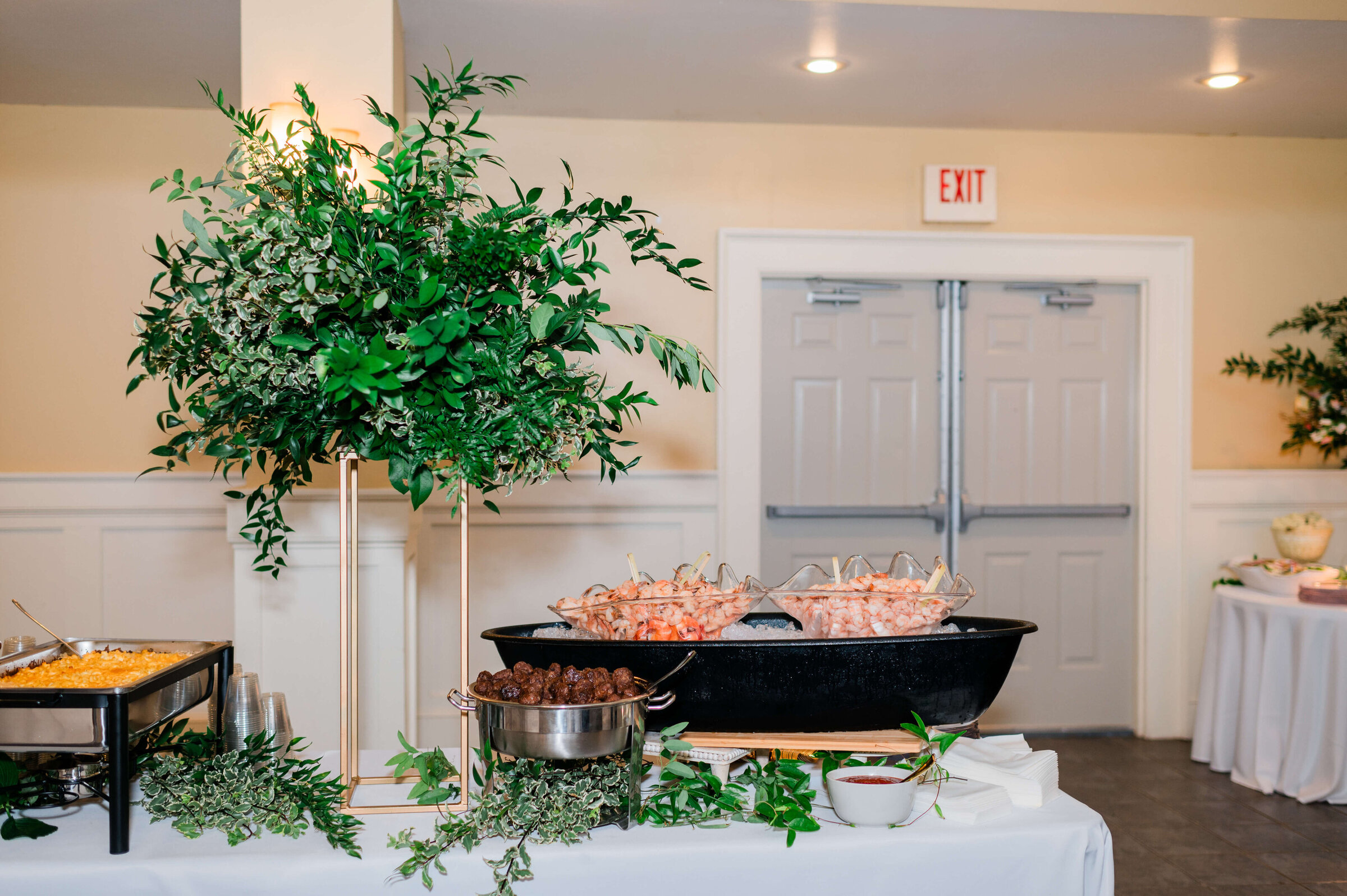 A buffet table with a large platter of shrimp on ice, a bowl of meatballs, and pans of hot food, decorated with greenery and a large floral arrangement, in a bright room.