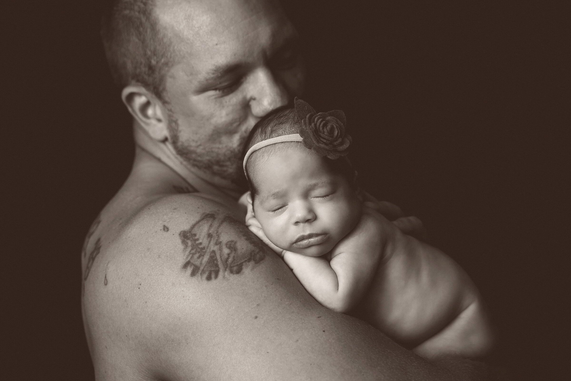 A shirtless man with tattoos on his arm holds a sleeping baby against his chest. The baby is wearing a headband with a small flower. The background is dark, highlighting the tender moment between the man and the baby, beautifully captured by a Jacksonville newborn photographer.