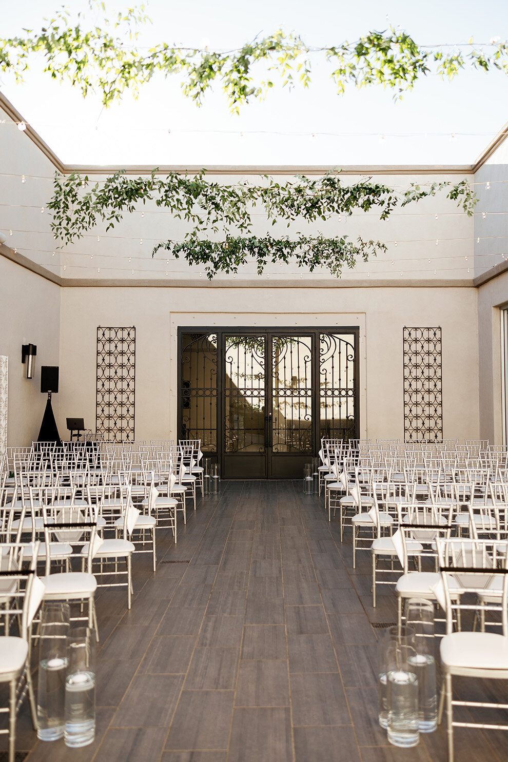 An elegant courtyard ceremony space at SoHo63, a Chandler wedding and event venue, is arranged with rows of silver chairs under a skylight ceiling. Greenery and string lights hang above the aisle, creating a modern and airy atmosphere perfect for weddings in downtown Chandler.