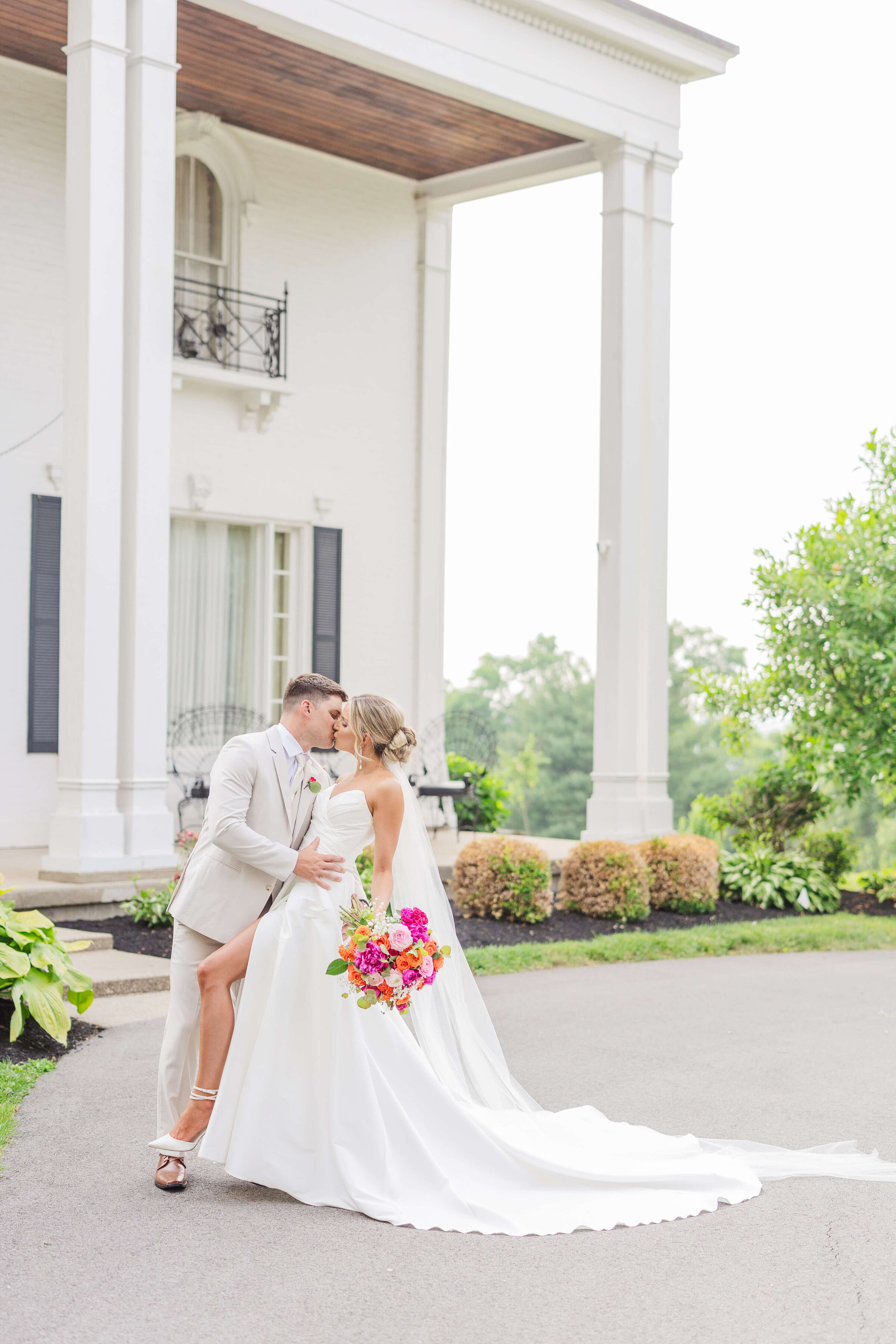 A groom in a tan suit dips his bride back for a kiss. They are standing in front of a white mansion outside. It's summertime and the trees are green and the sun is glowing. She's holding a bright pink bouquet.